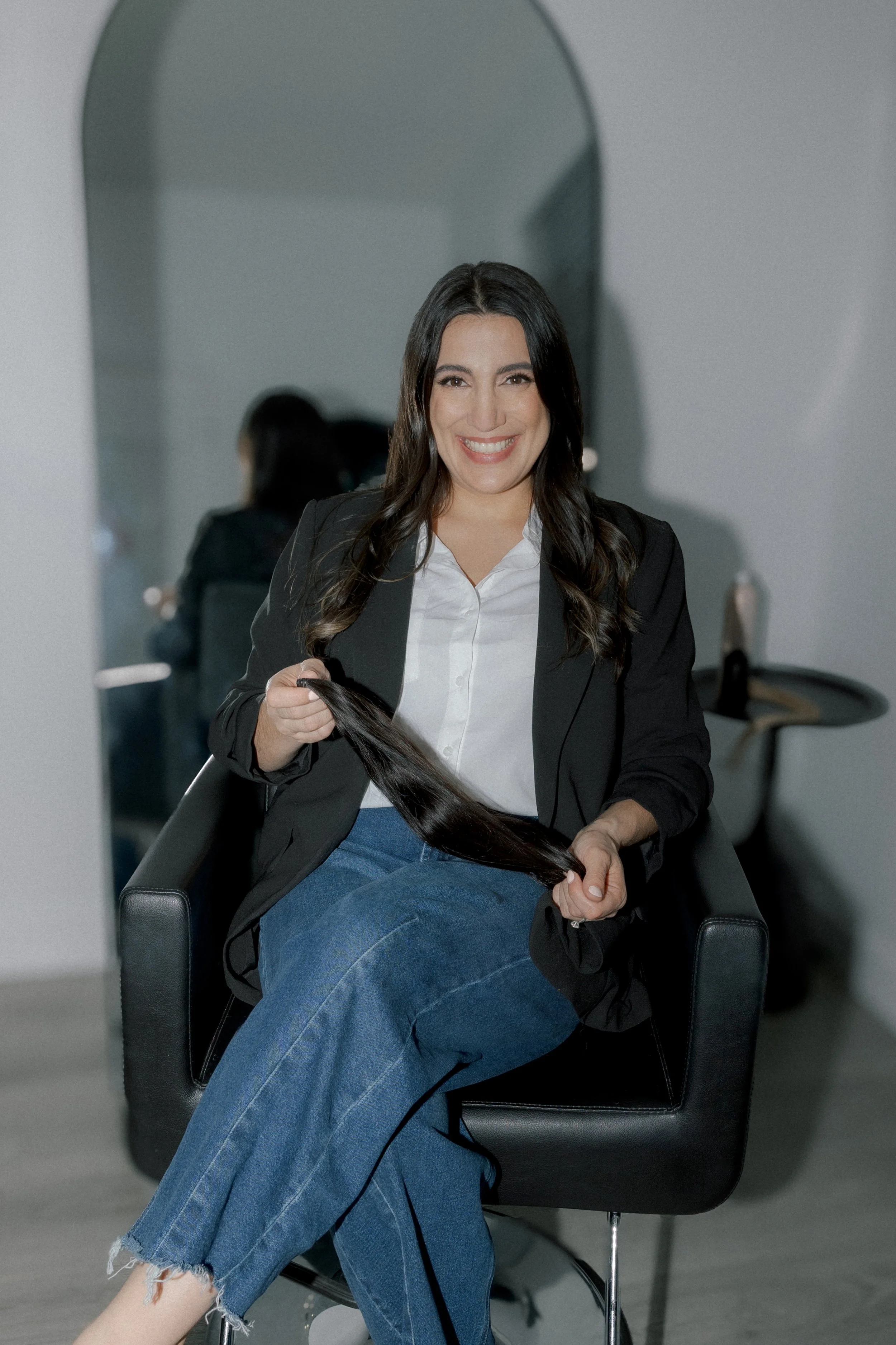 Woman sitting in a salon chair, holding a lock of long, dark hair, smiling at the camera.