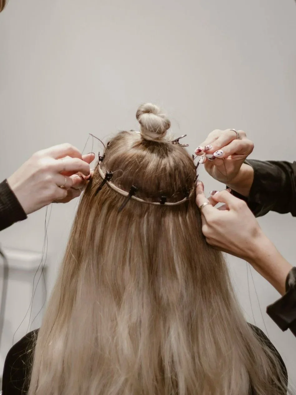 Two people are weaving hair extensions into a woman's hair, with her head turned away. The woman has blonde hair, and the background is neutral.