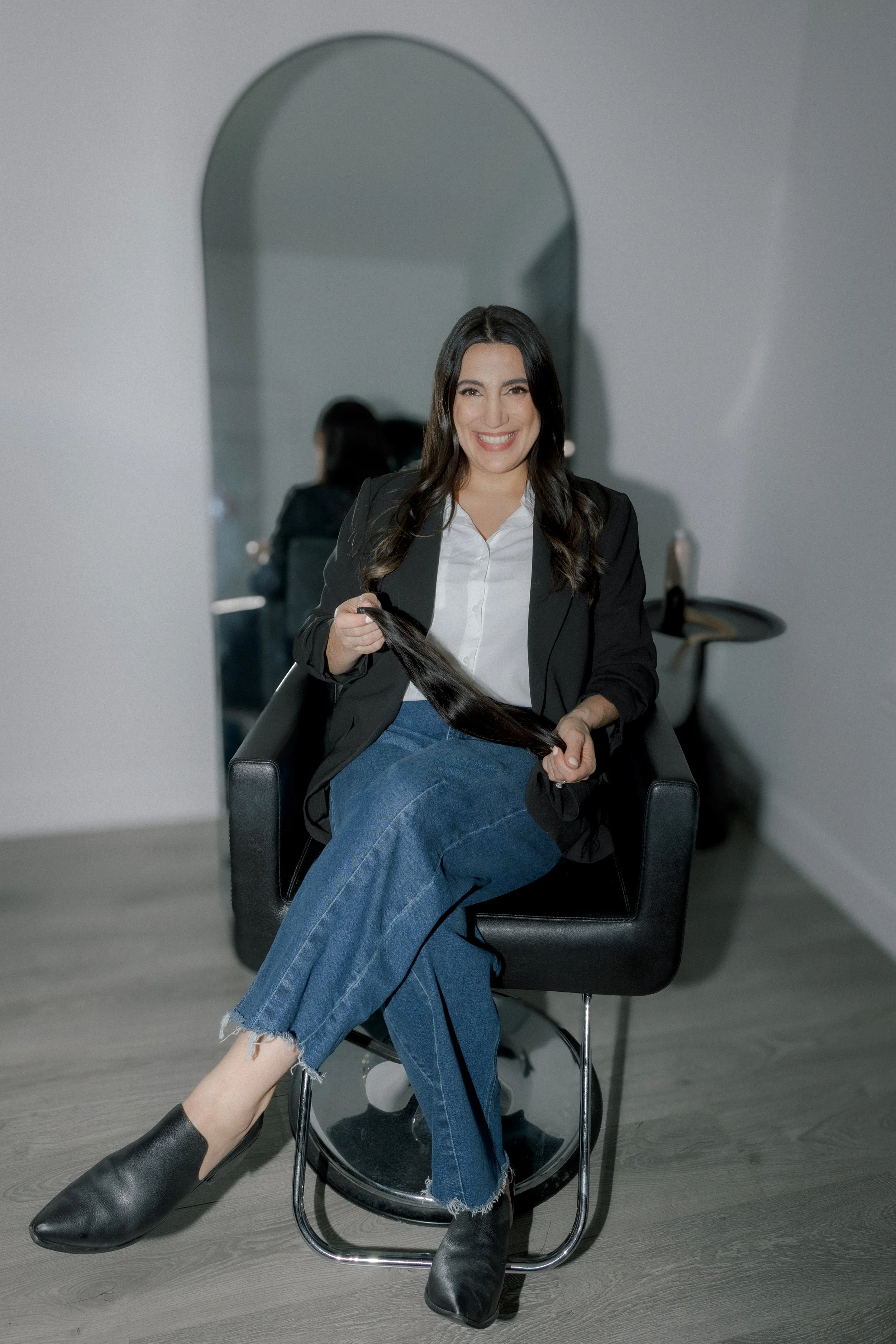 Woman sitting in salon chair smiling, holding long dark hair, in front of mirror.