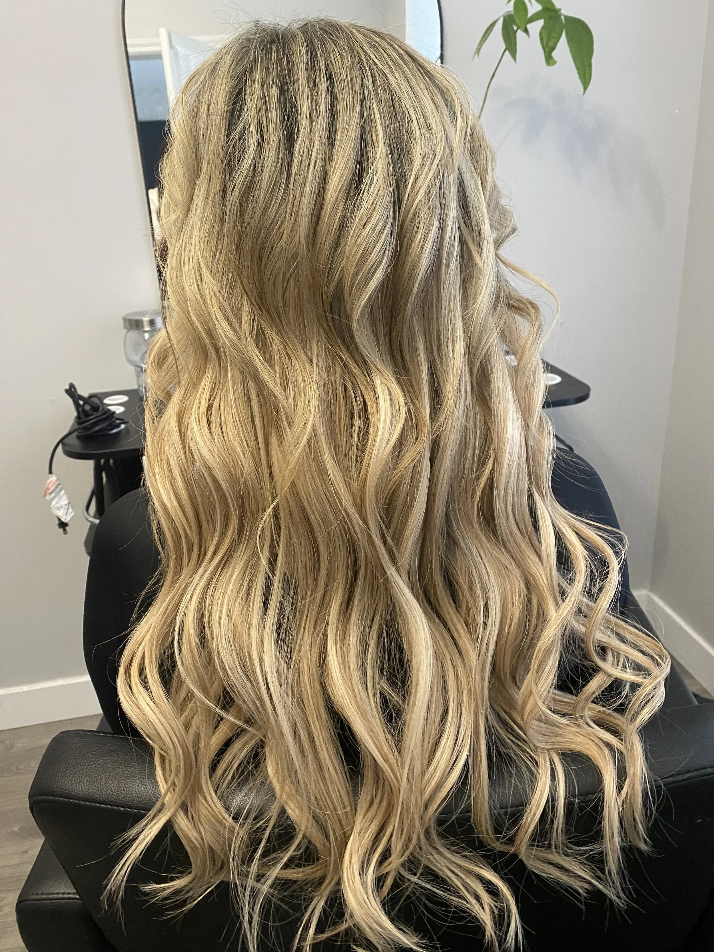 Back view of a woman with long, blonde, wavy hair sitting in a stylist chair in a salon.