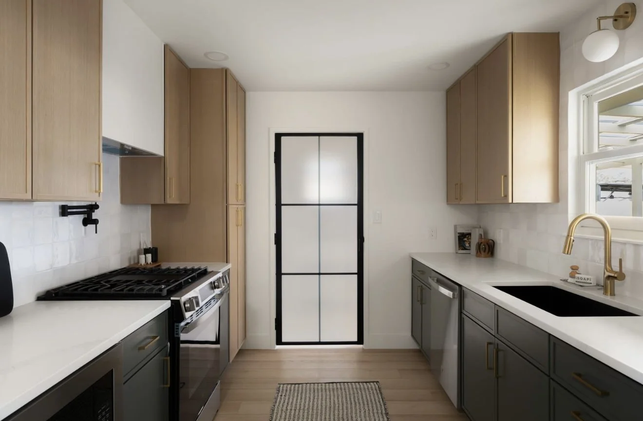 Modern kitchen with white countertops, wooden upper cabinets, gray lower cabinets, black sink, gold hardware, and a black-framed glass door.