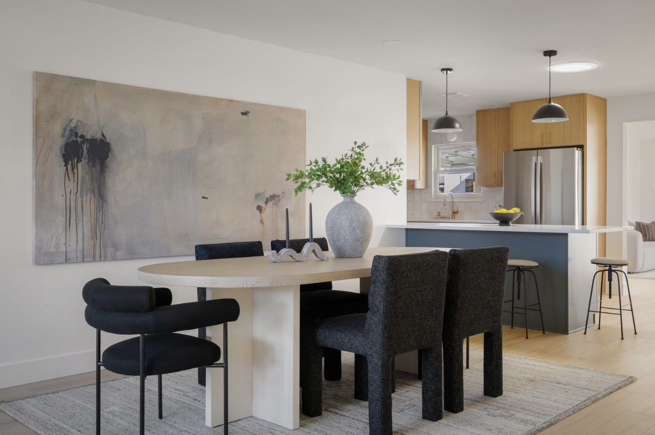 Modern dining area with a wooden table, black chairs, a large abstract painting on the wall, and a kitchen with wooden cabinets, stainless steel refrigerator, and black pendant lights.