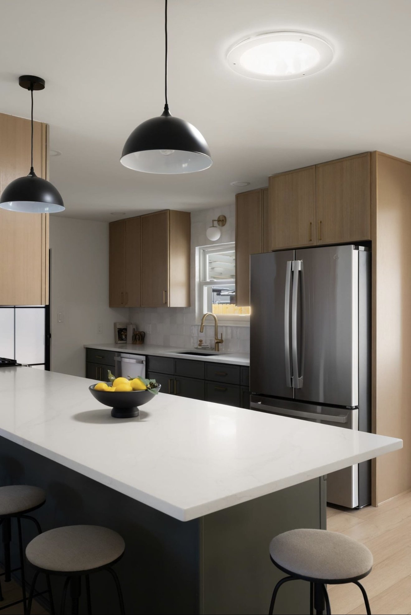Modern kitchen with black pendant lights over a white island, wooden upper cabinets, black lower cabinets, stainless steel refrigerator, and a window with a gold faucet
