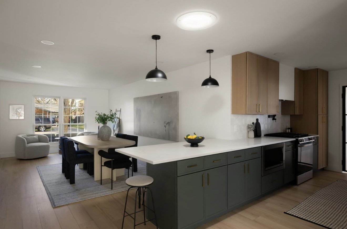 Open kitchen and dining area with a white island, green cabinets, wooden upper cabinets, and black appliances; dining table with black and beige chairs, a vase with green foliage; living space with white armchair and large window.