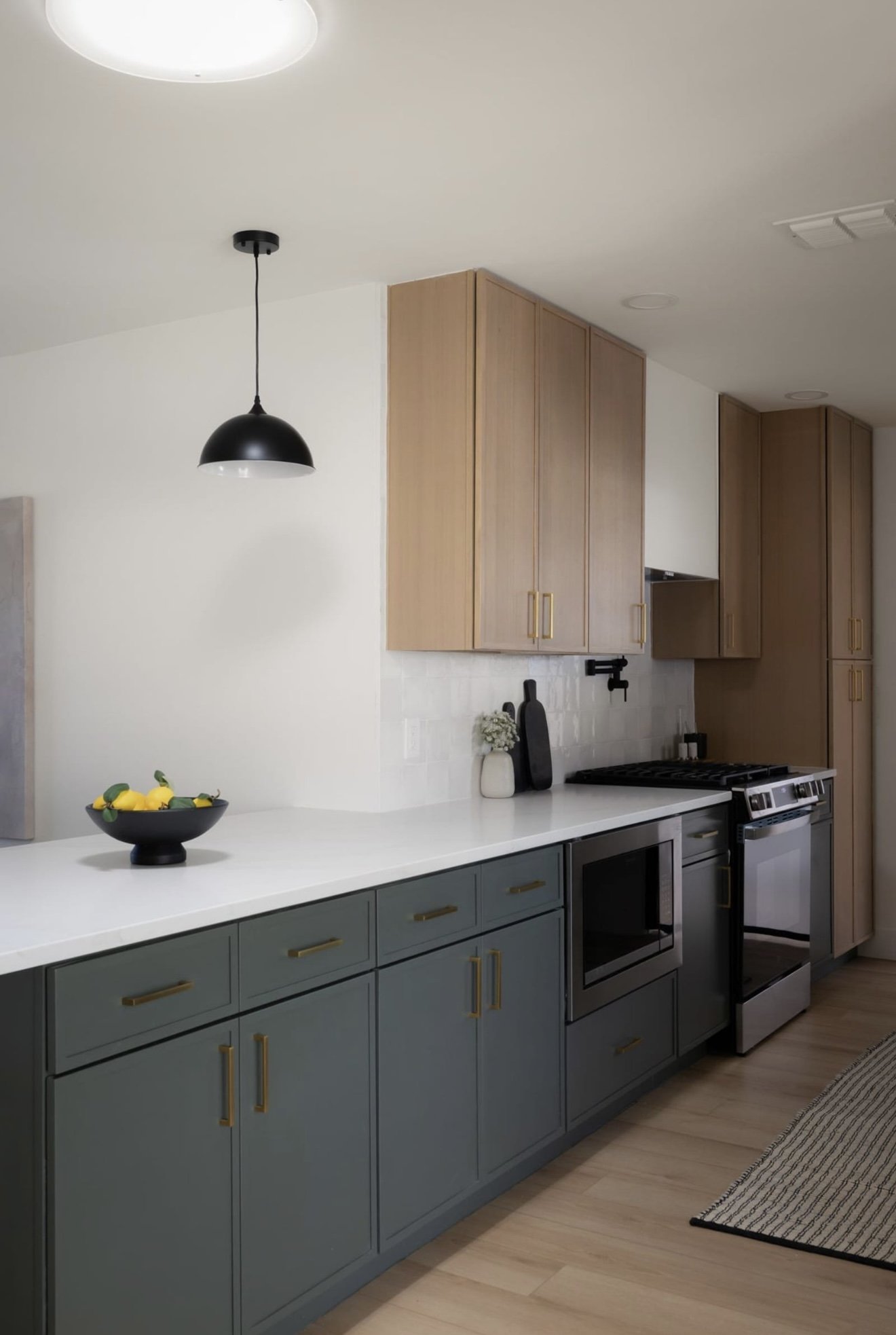 Modern kitchen with sage green lower cabinets, natural wood upper cabinets, white countertop, black pendant light, stainless steel stove and microwave, decorative vases, black cutting boards, lemon bowl, and a striped rug on light wood floors.