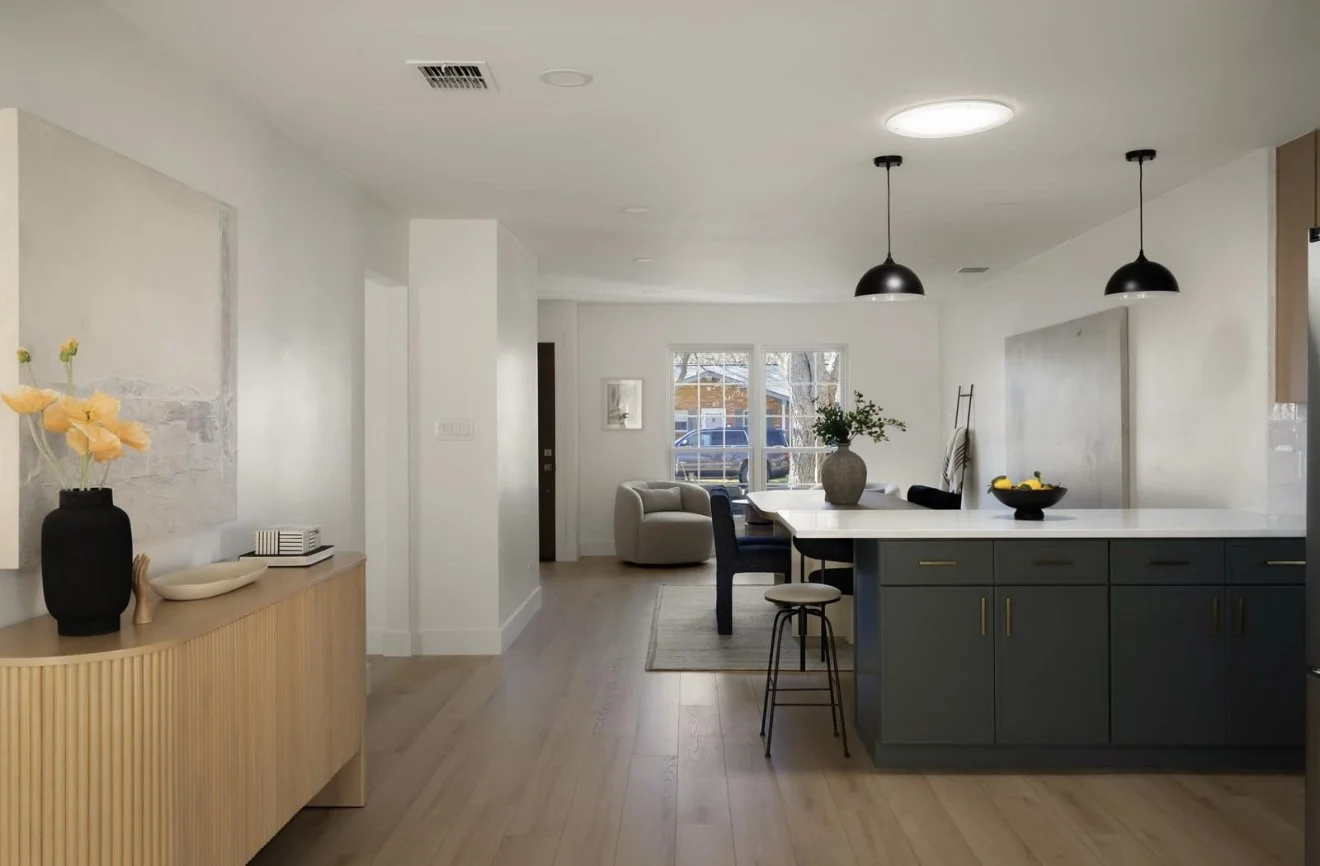 Open concept living and dining area with a kitchen featuring a dark green island, modern black pendant lights, a white table with a gray vase, and a cozy seating area by the window with natural light.