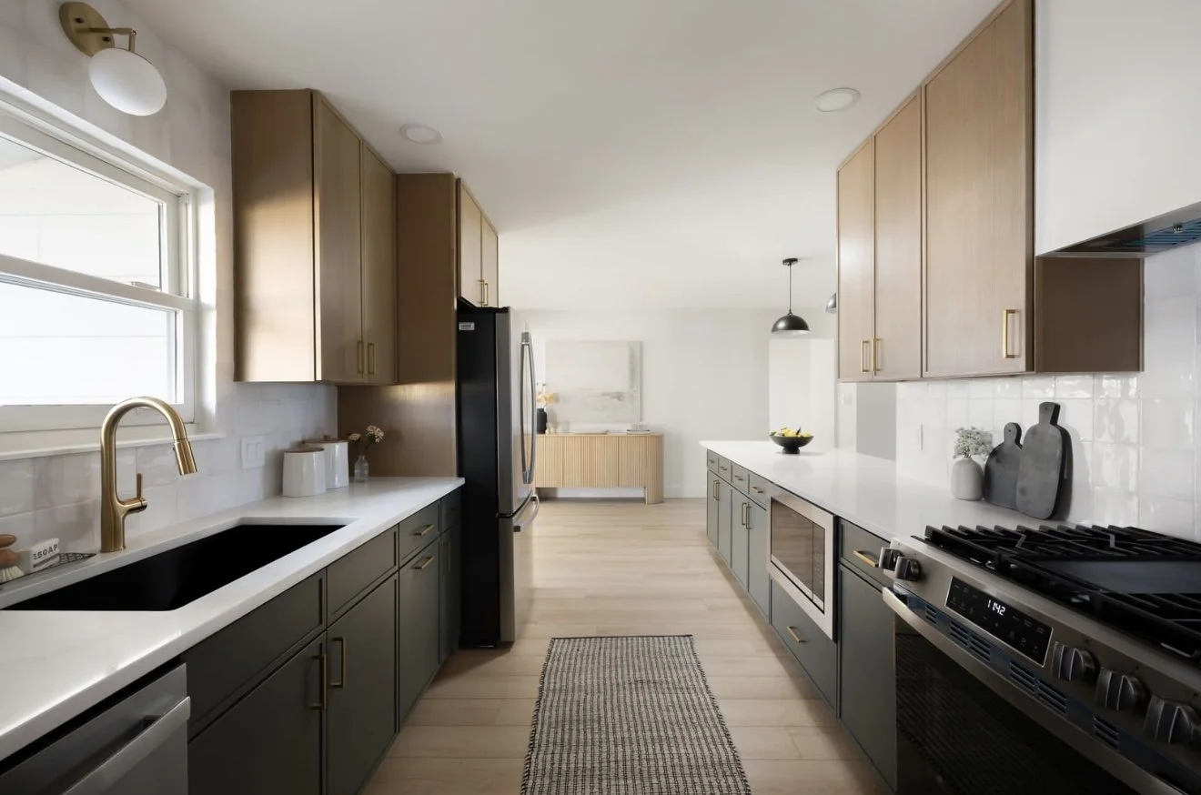 Modern kitchen with black sink, gold faucet, wooden cabinets, white countertops, stainless steel refrigerator, oven, and stove, with a window above the sink, a pendant light, and decorative items including cutting boards and vases.
