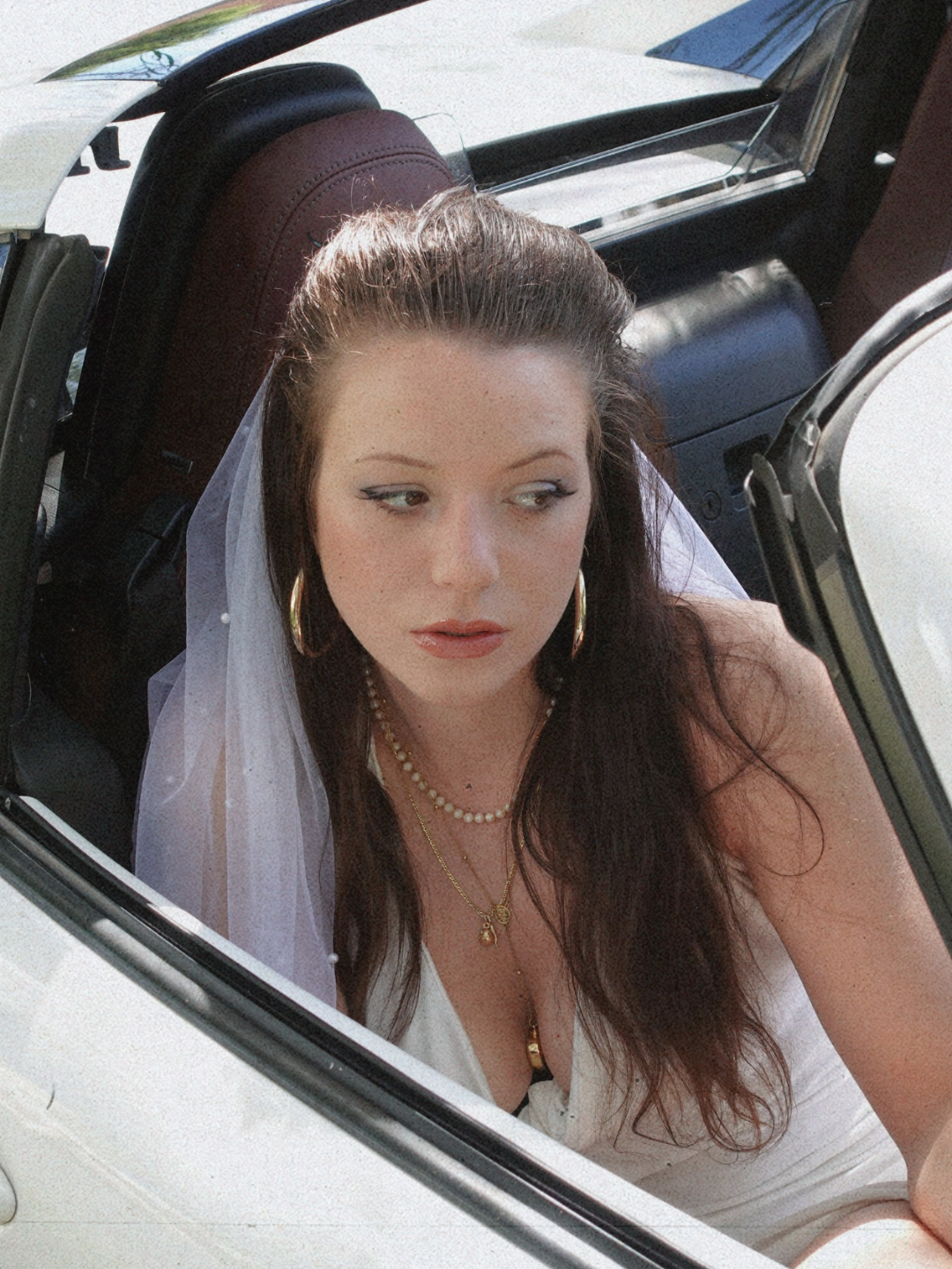 A woman with long dark hair, wearing hoop earrings, necklaces, and a white outfit, sitting in the driver's seat of a vintage convertible car. She looks to the side through the open window.
