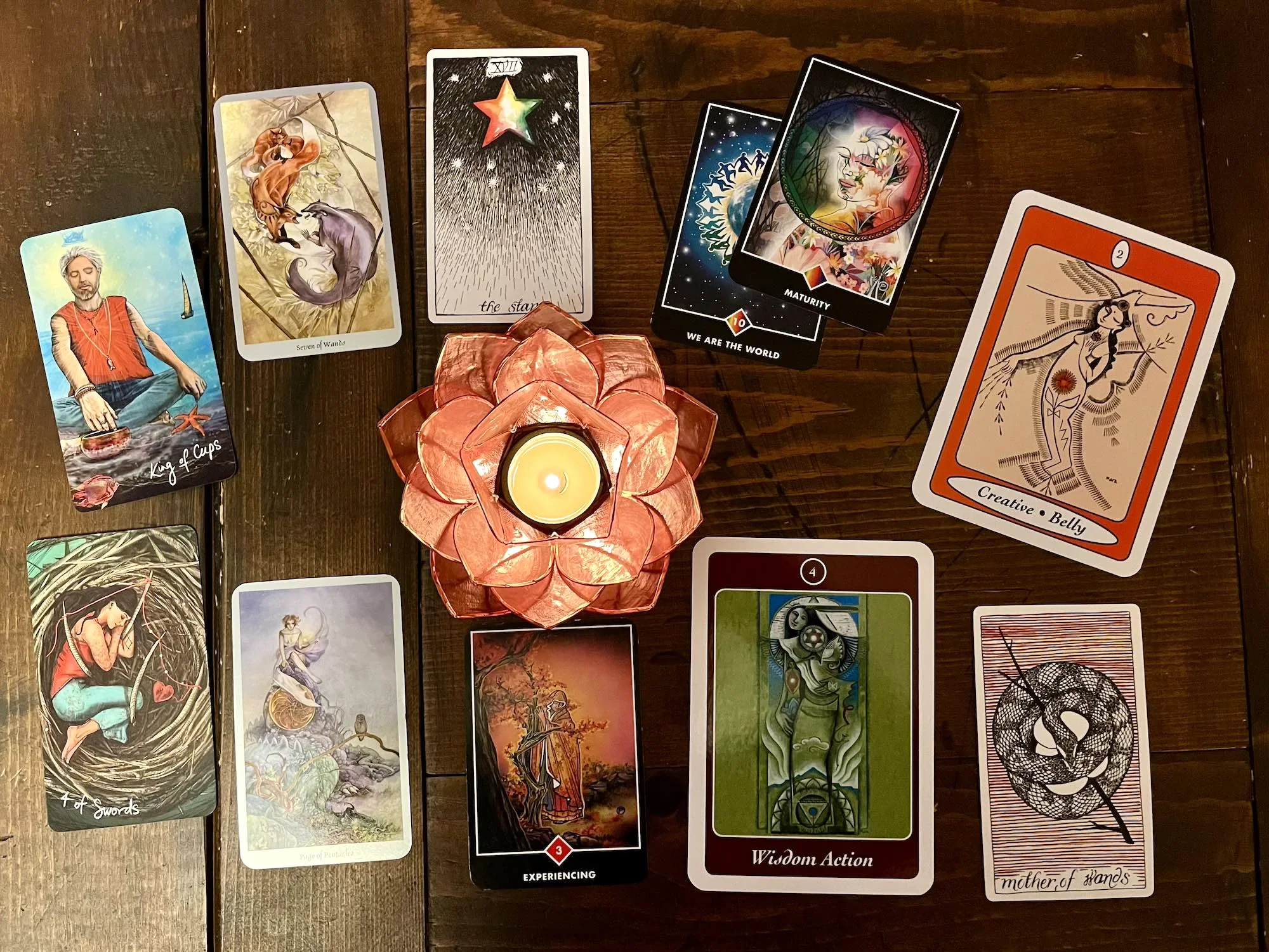 A table with tarot cards laid out, including the King of Cups, Seven of Wands, the Star, and other cards with abstract and artistic designs, surrounding a lit candle in a decorative holder.