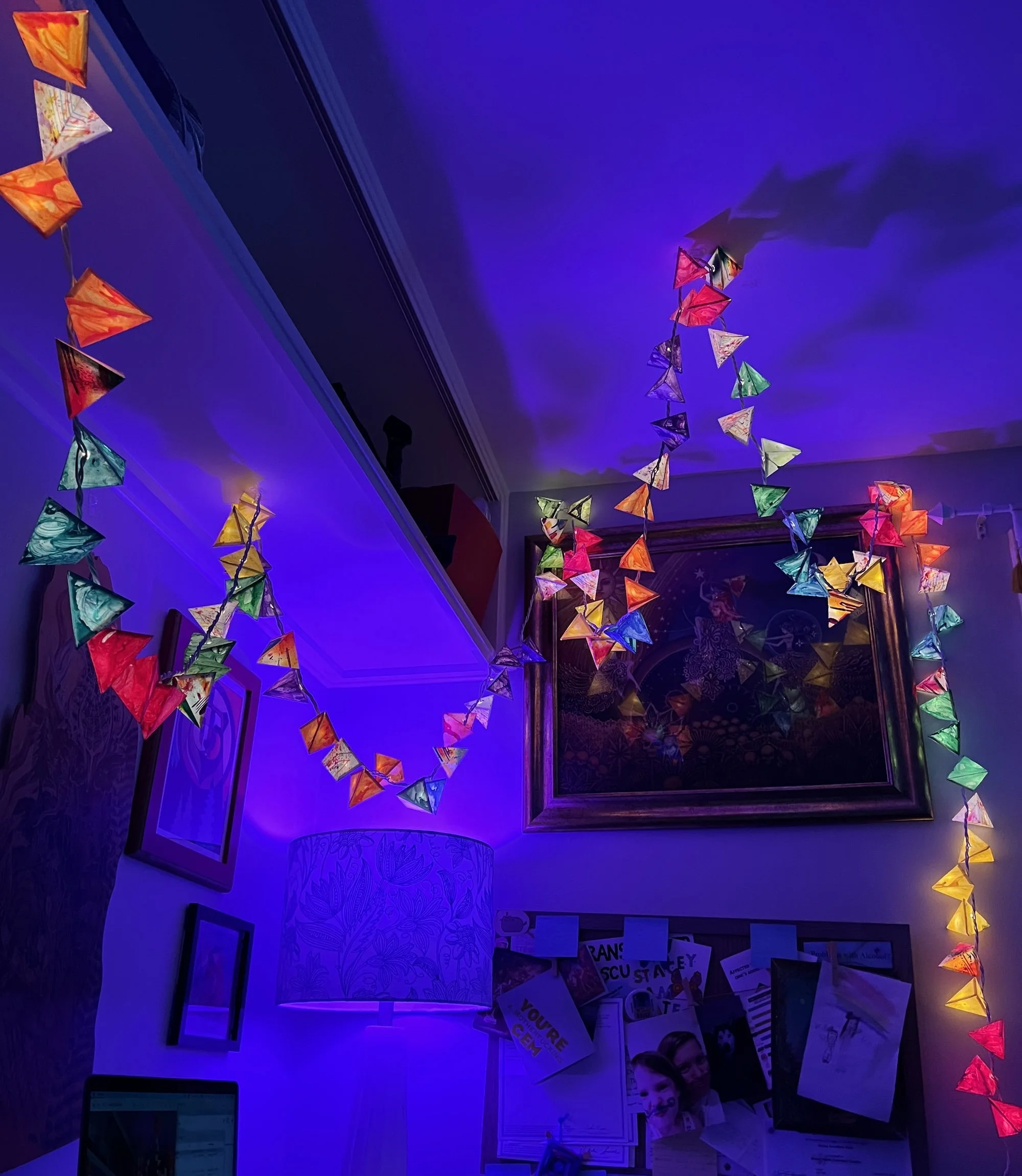Colorful triangular string lights draped across a wall and ceiling, glowing against purple ambient lighting above framed artwork, a lamp, and a bulletin board.