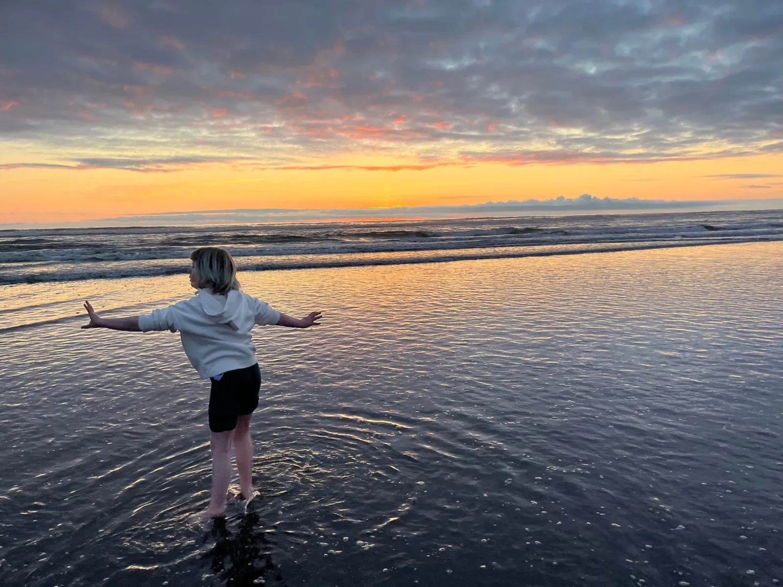 A child with blonde hair, wearing a white hoodie and black shorts, standing in shallow ocean water at sunset with arms outstretched and looking towards the horizon