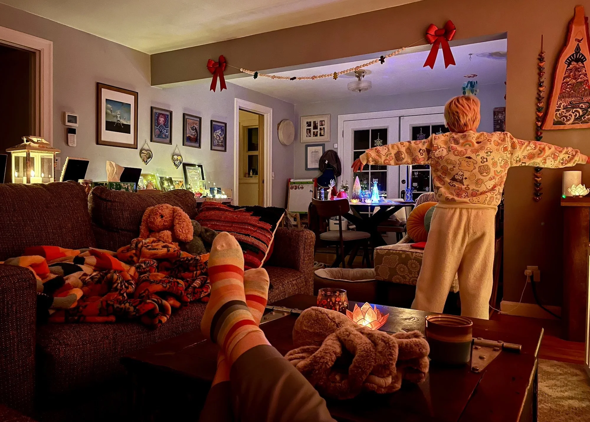 Cozy living room lit with warm lamps and candles; a child stands with arms outstretched near a table of glowing decorations, while socked feet rest on a couch in the foreground among blankets and stuffed animals.