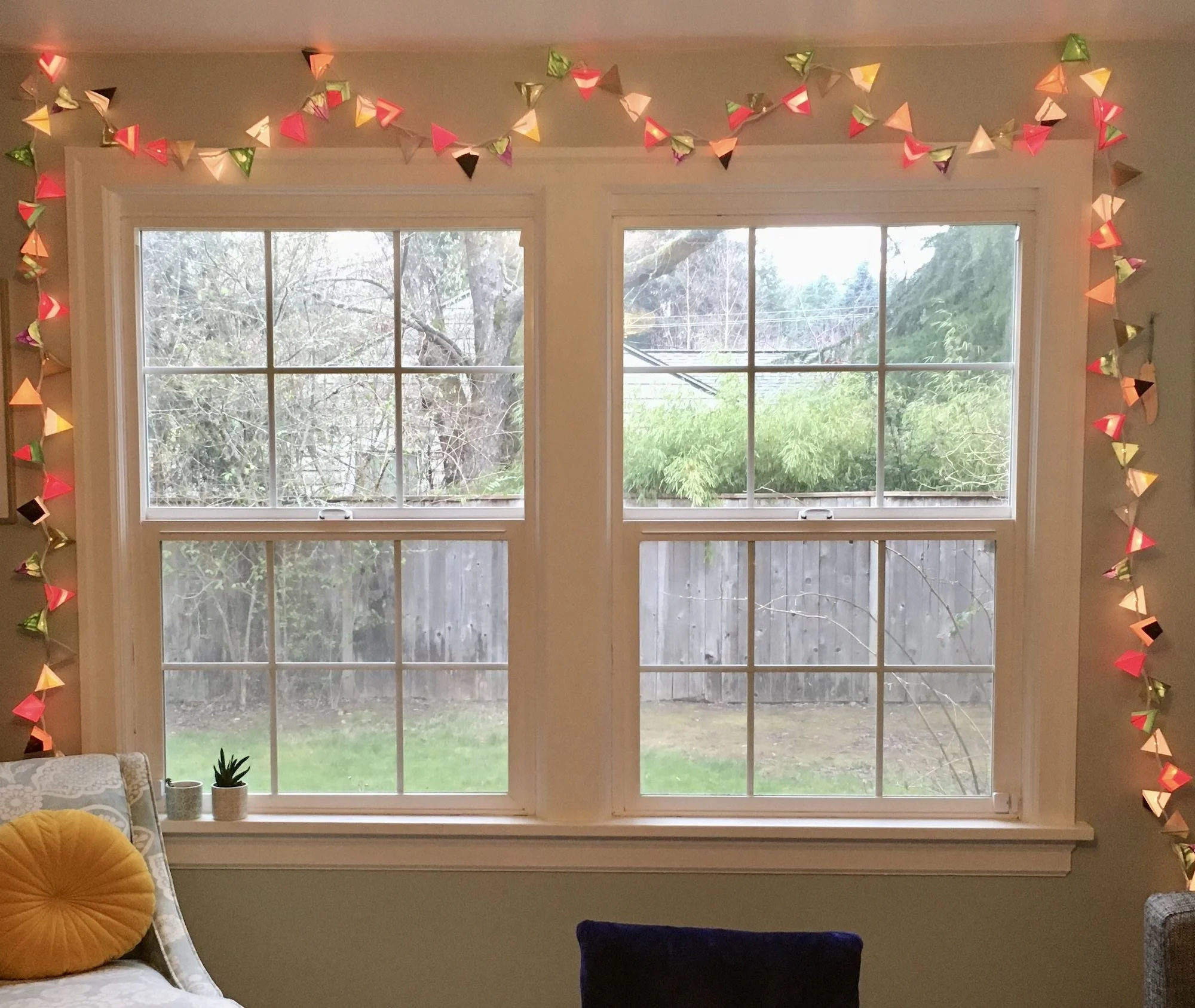 Two side-by-side windows framed with a string of colorful triangular lights, looking out onto a green backyard; soft pillows and a small plant sit below the windows.