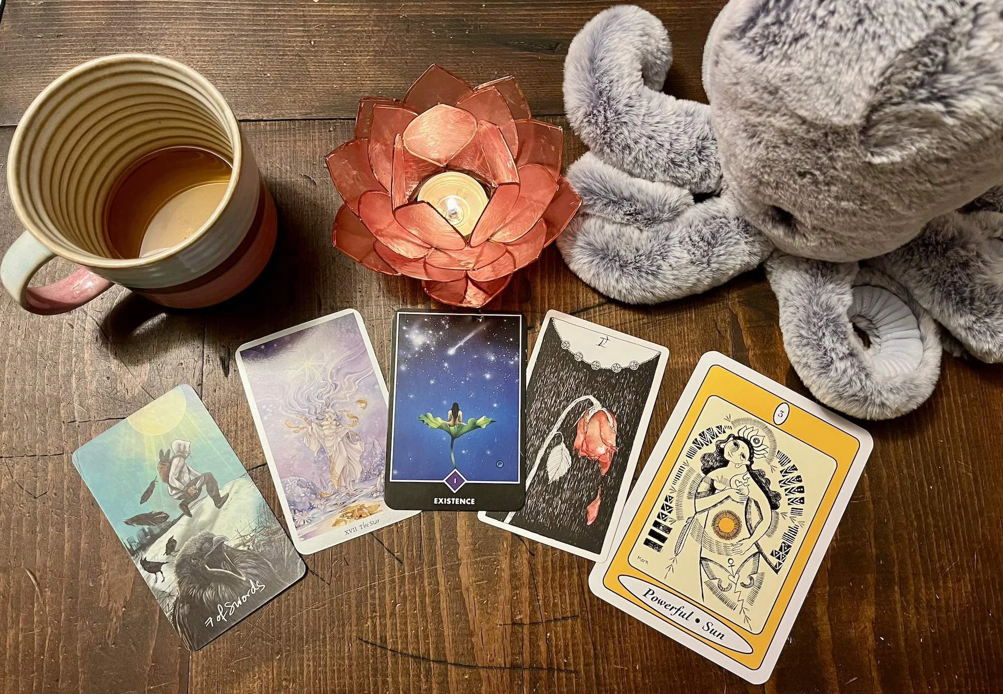 Overhead view of a wooden table with a tarot spread arranged in a row beneath a lit candle in a pink lotus-shaped holder, alongside a ceramic mug and a gray plush octopus.