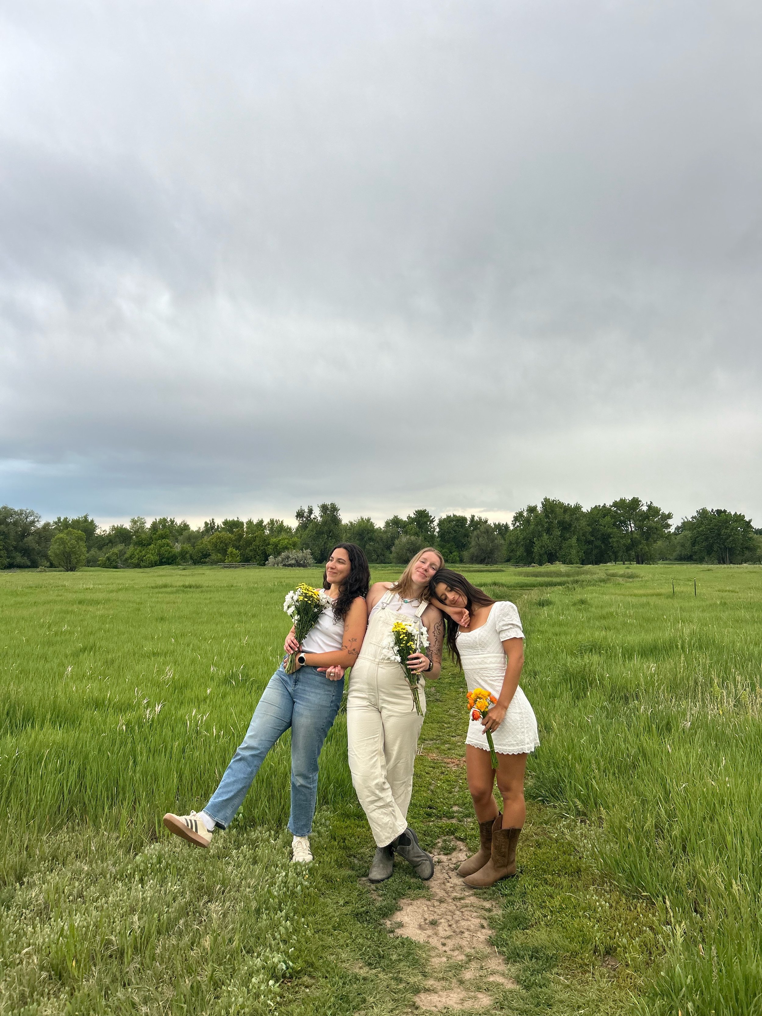 Three women standing on a dirt path in a green field, each holding a bouquet of flowers, with trees and a cloudy sky in the background.