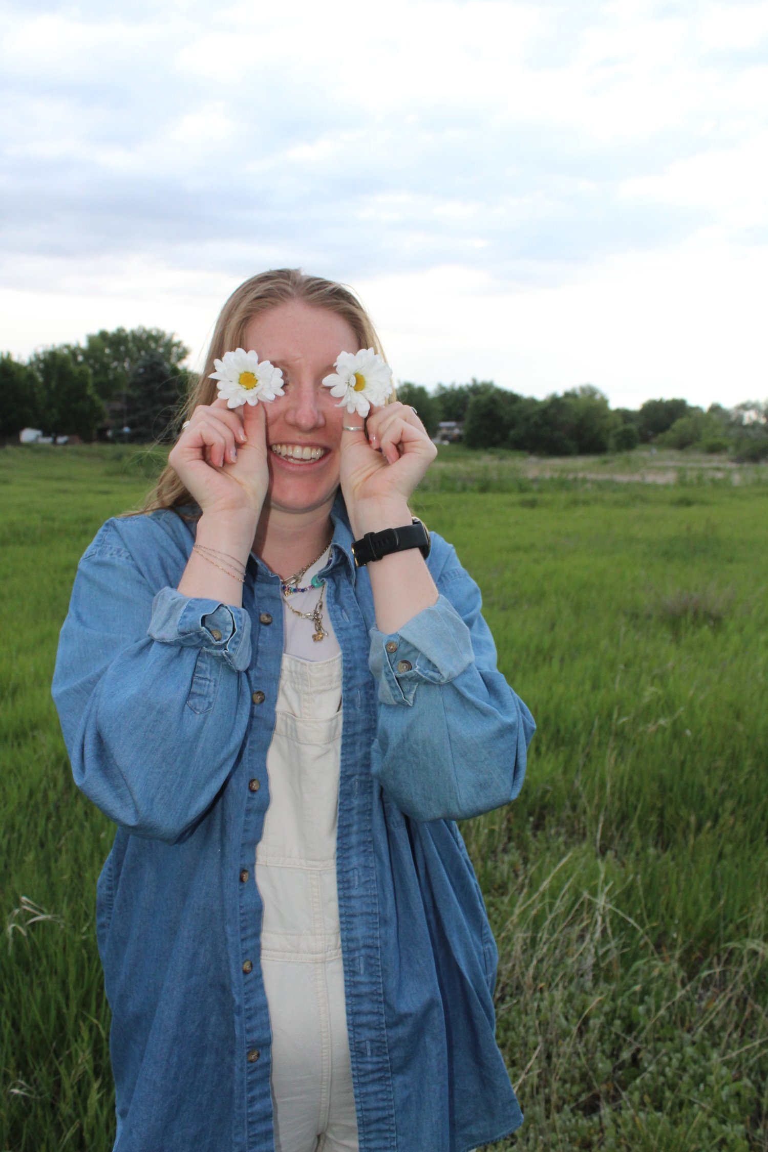Woman smiling in a grassy field, holding daisies in front of her eyes