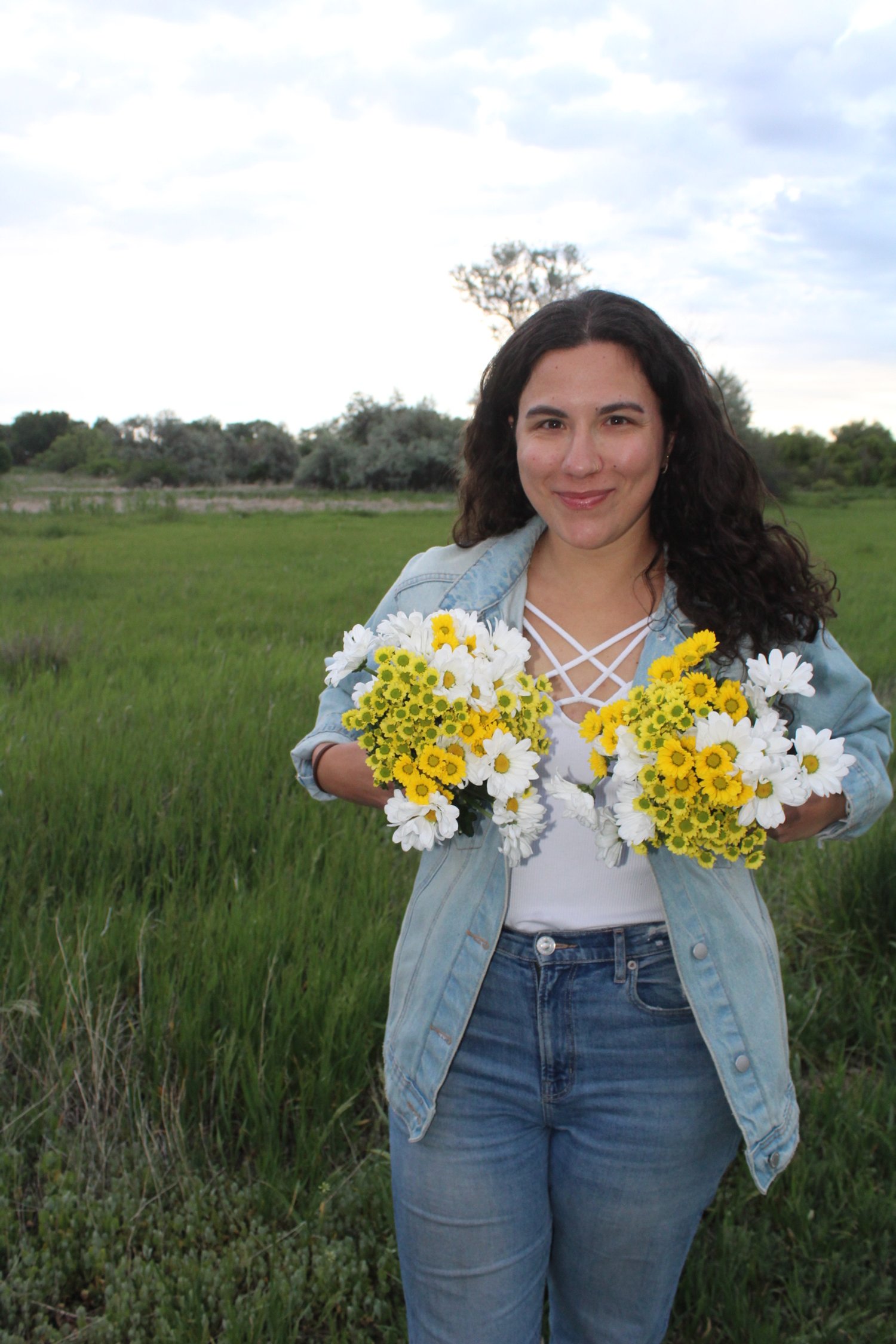 Young woman standing outdoors on a grassy field, holding two bouquets of white and yellow daisies, smiling at the camera, with trees and a cloudy sky in the background.