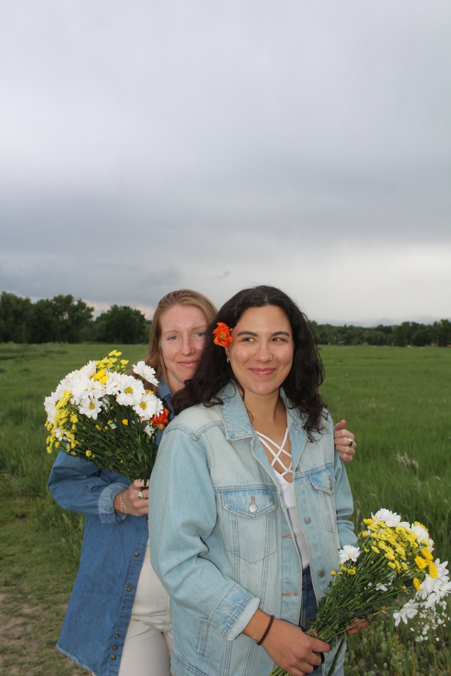 Two women standing outdoors in a grassy field holding bouquets of flowers, with cloudy sky in the background.