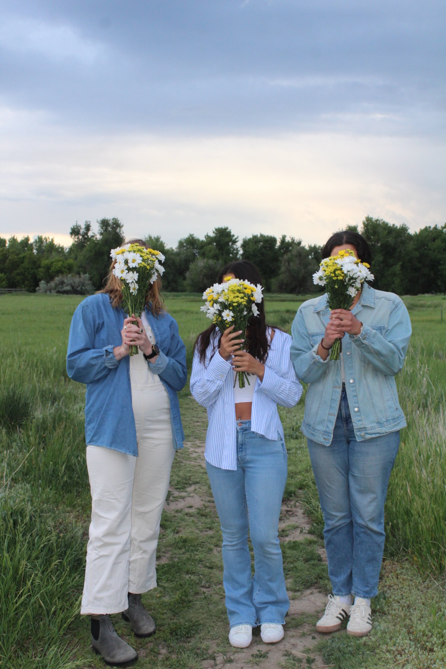 Three women standing outdoors in a grassy area holding bouquets of white and yellow flowers in front of their faces.