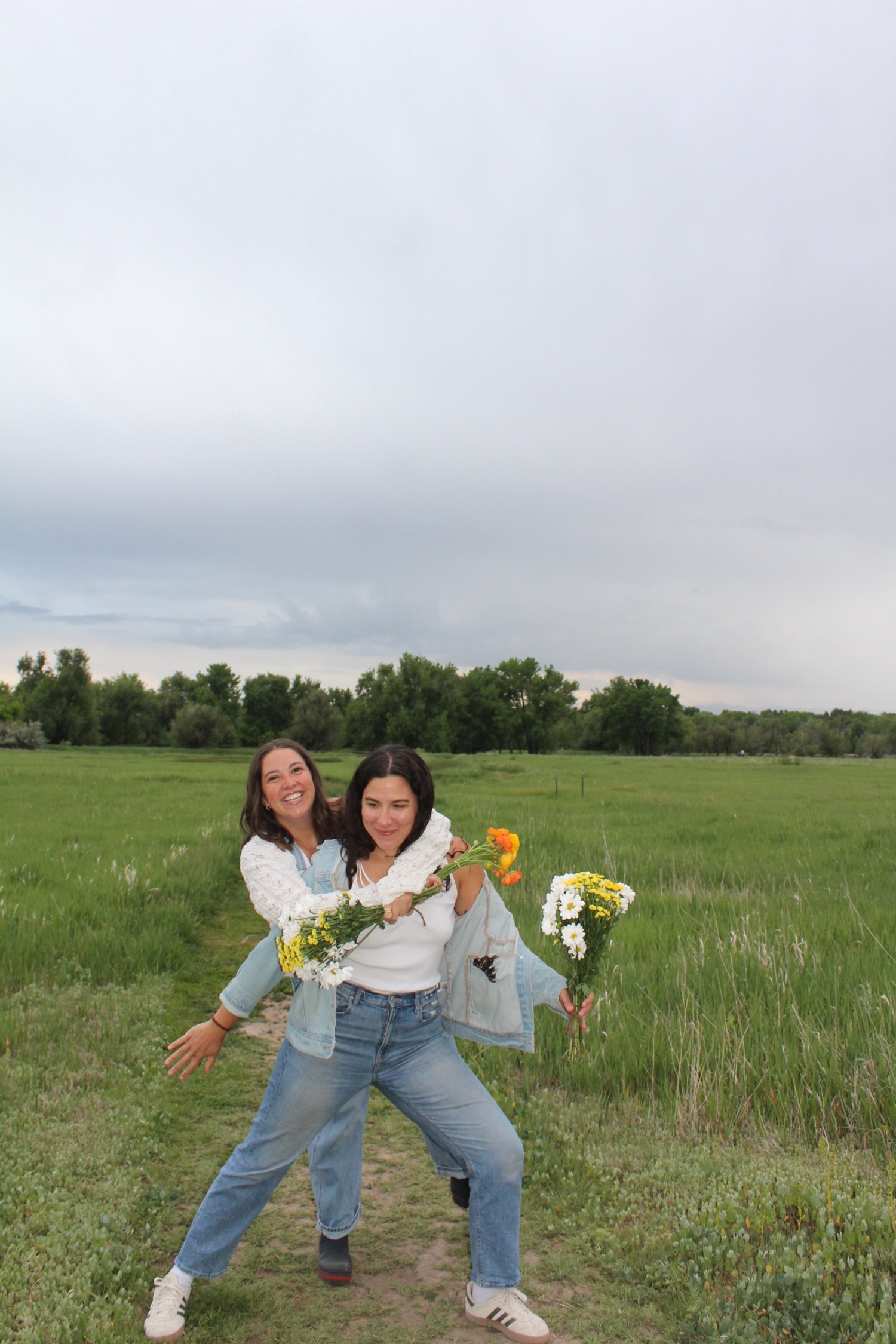 Two young women standing on a grassy path in a green field with trees in the background, holding bouquets of flowers and sharing a joyful moment outdoors under a cloudy sky.