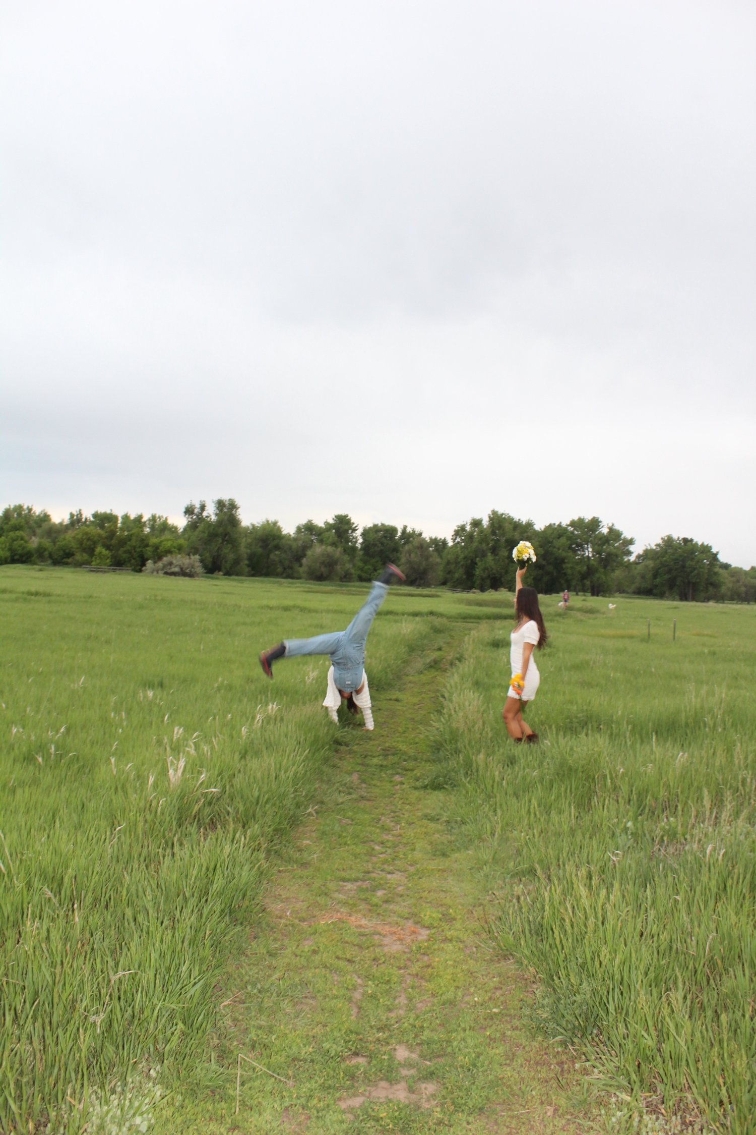 A woman in a white dress holding a bouquet standing on a grassy path, while another person in casual clothes is doing a cartwheel nearby in an open field with trees in the background and an overcast sky.