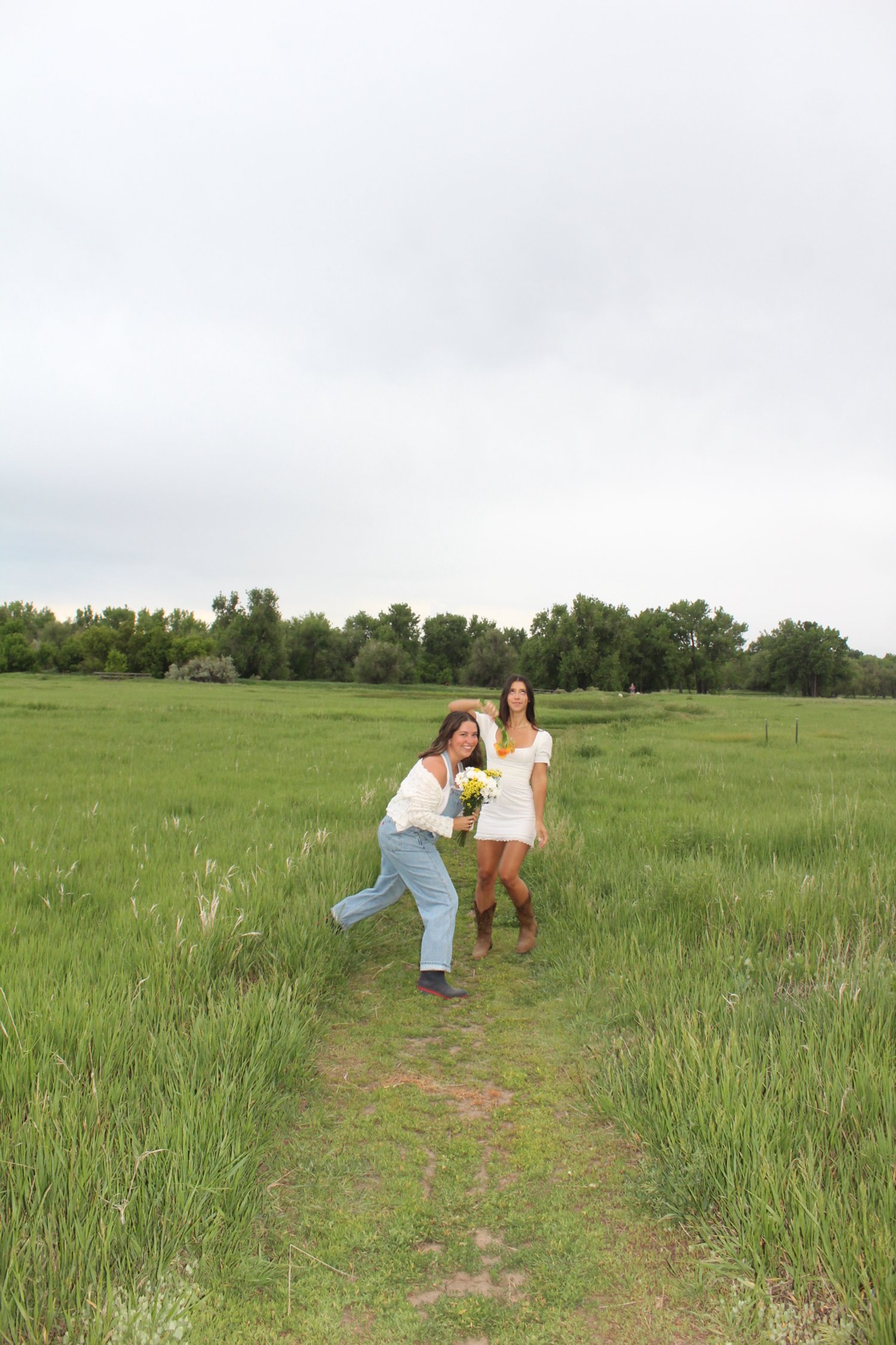 Two women standing in a green field with trees in the background. One woman is holding a bouquet of white flowers, and the other woman is wearing a white dress and brown boots.