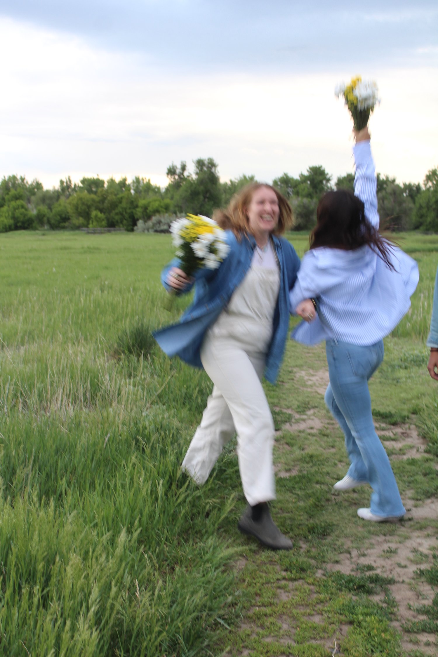 Two women joyfully celebrating outdoors with flowers, one woman holding flowers above her head and the other with flowers in her hand, on a grassy path with trees in the background.