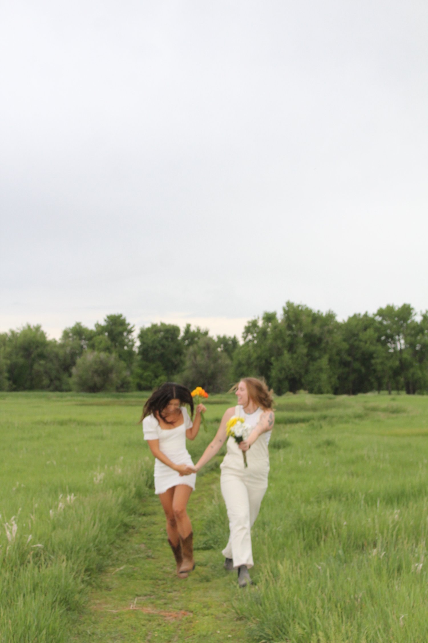 Two women walking on a grassy field holding flowers, one wearing a white dress and cowboy boots, the other in a cream jumpsuit, with trees and an overcast sky in the background.