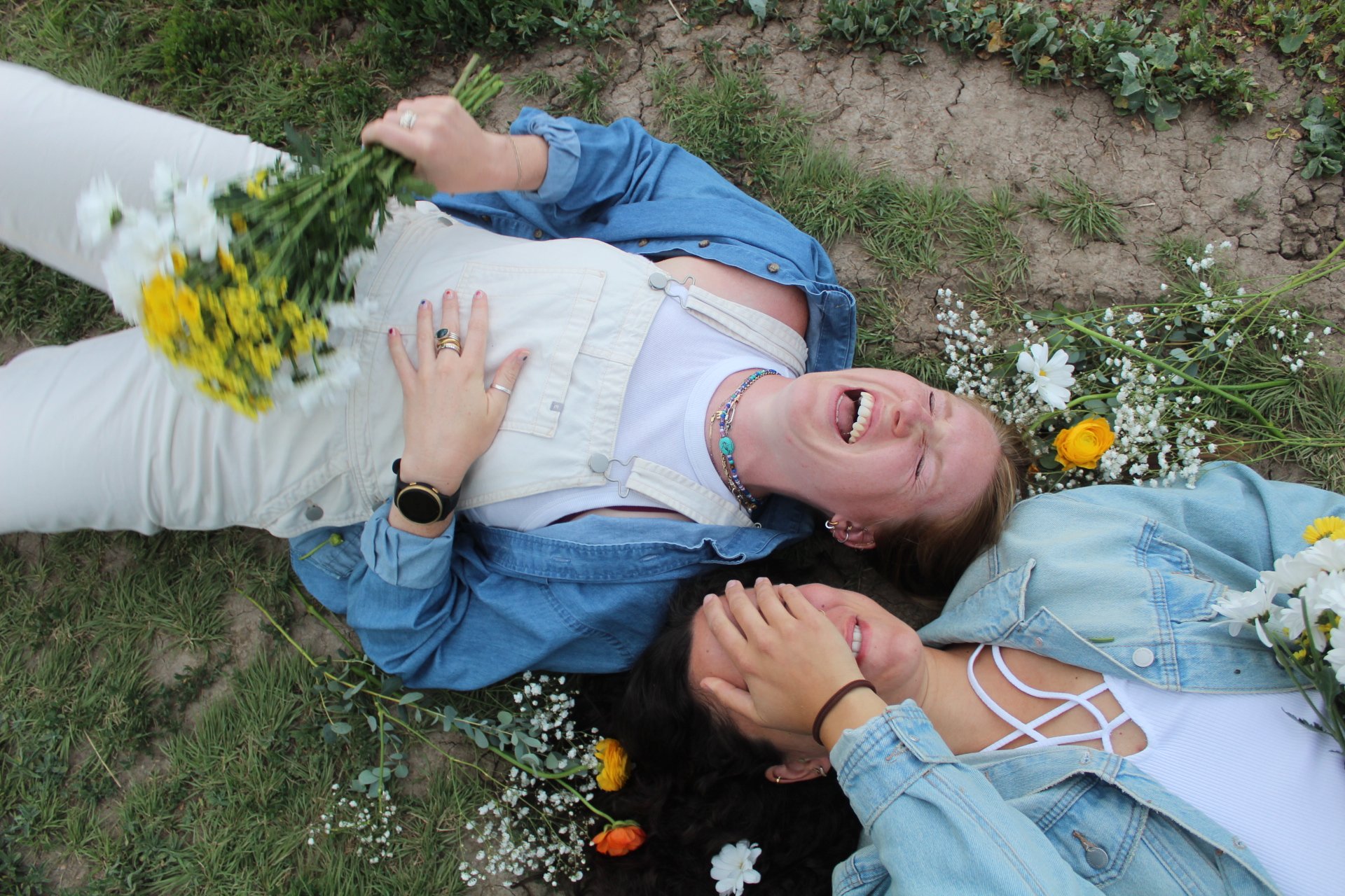 Two women lying on the grass, surrounded by flowers, laughing and enjoying each other's company.