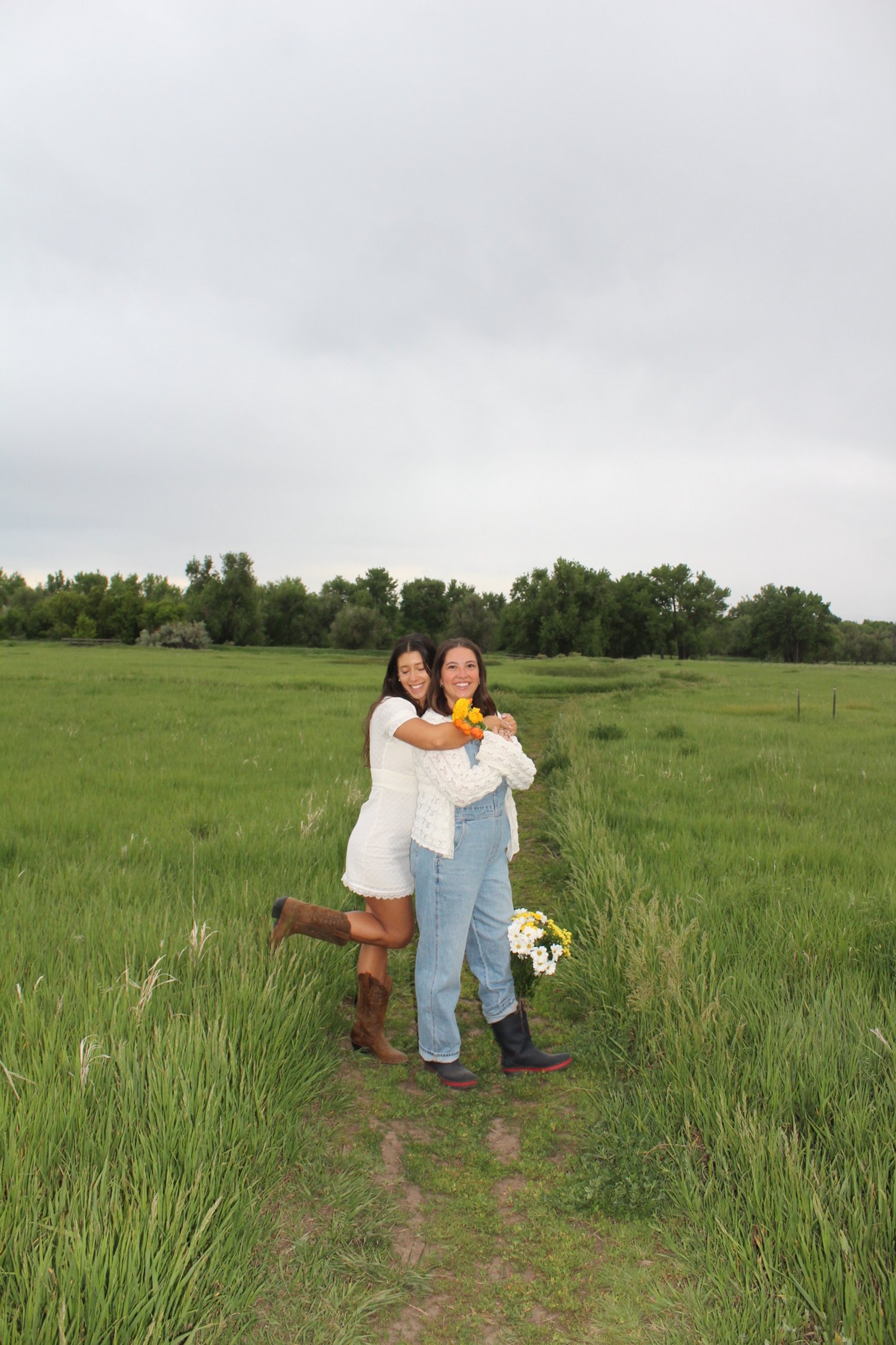Two women hugging on a grassy path in a field, one holding a bouquet of flowers, overcast sky.