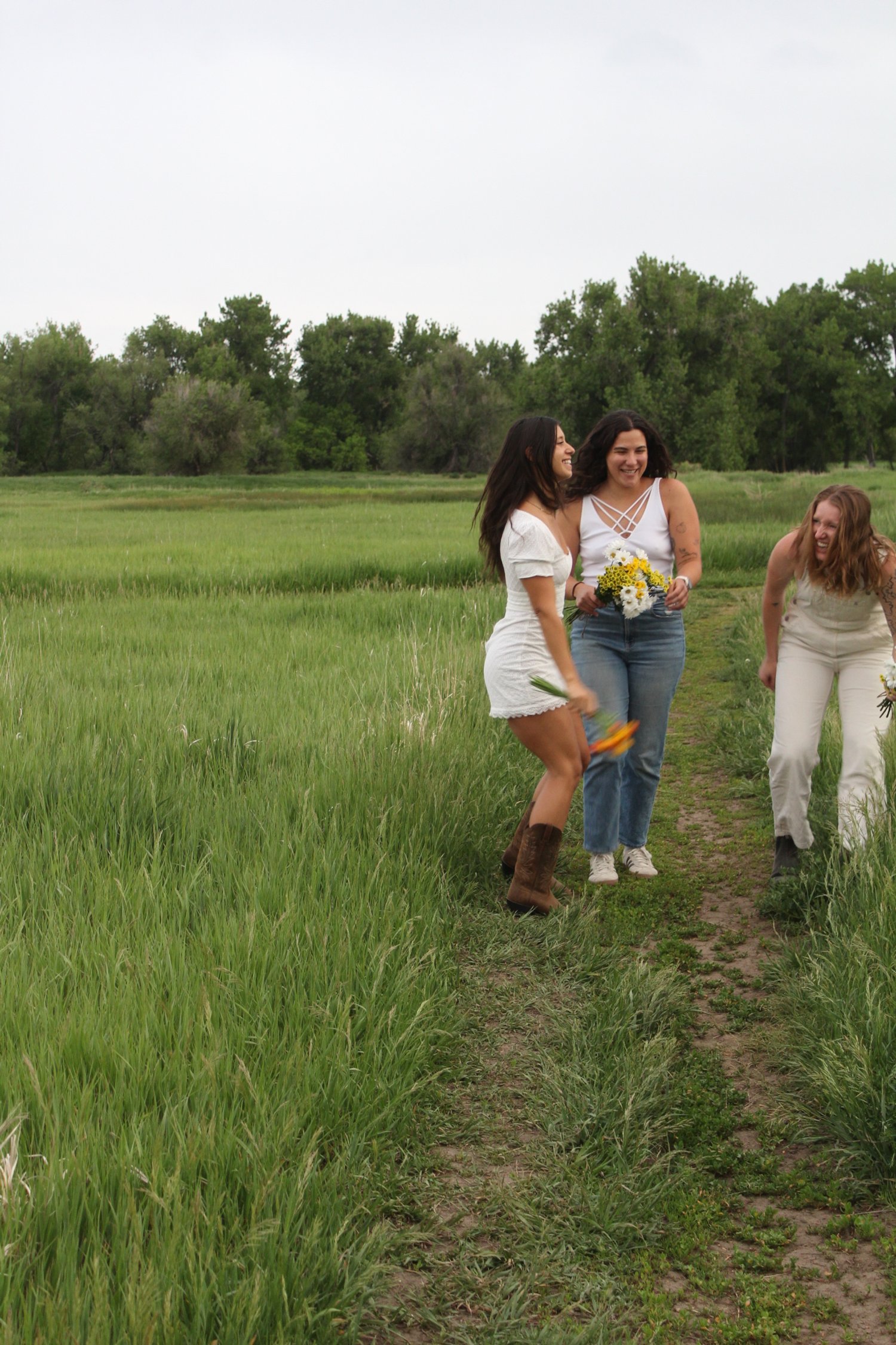 Three women in a green field, one holding flowers, laughing and talking during a cloudy day.