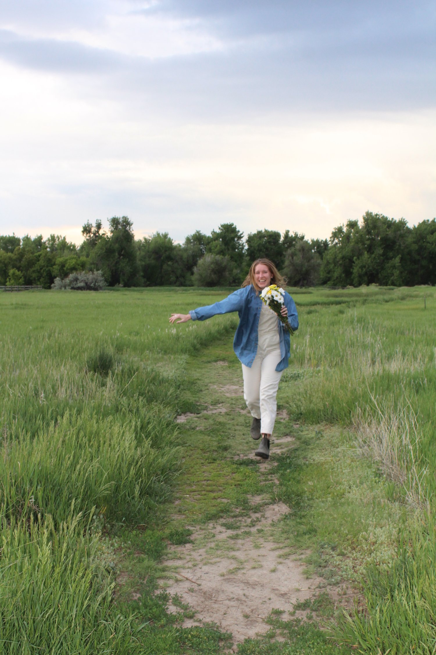 A woman running joyfully along a grassy path holding a bouquet of flowers in a lush, green field under a cloudy sky.