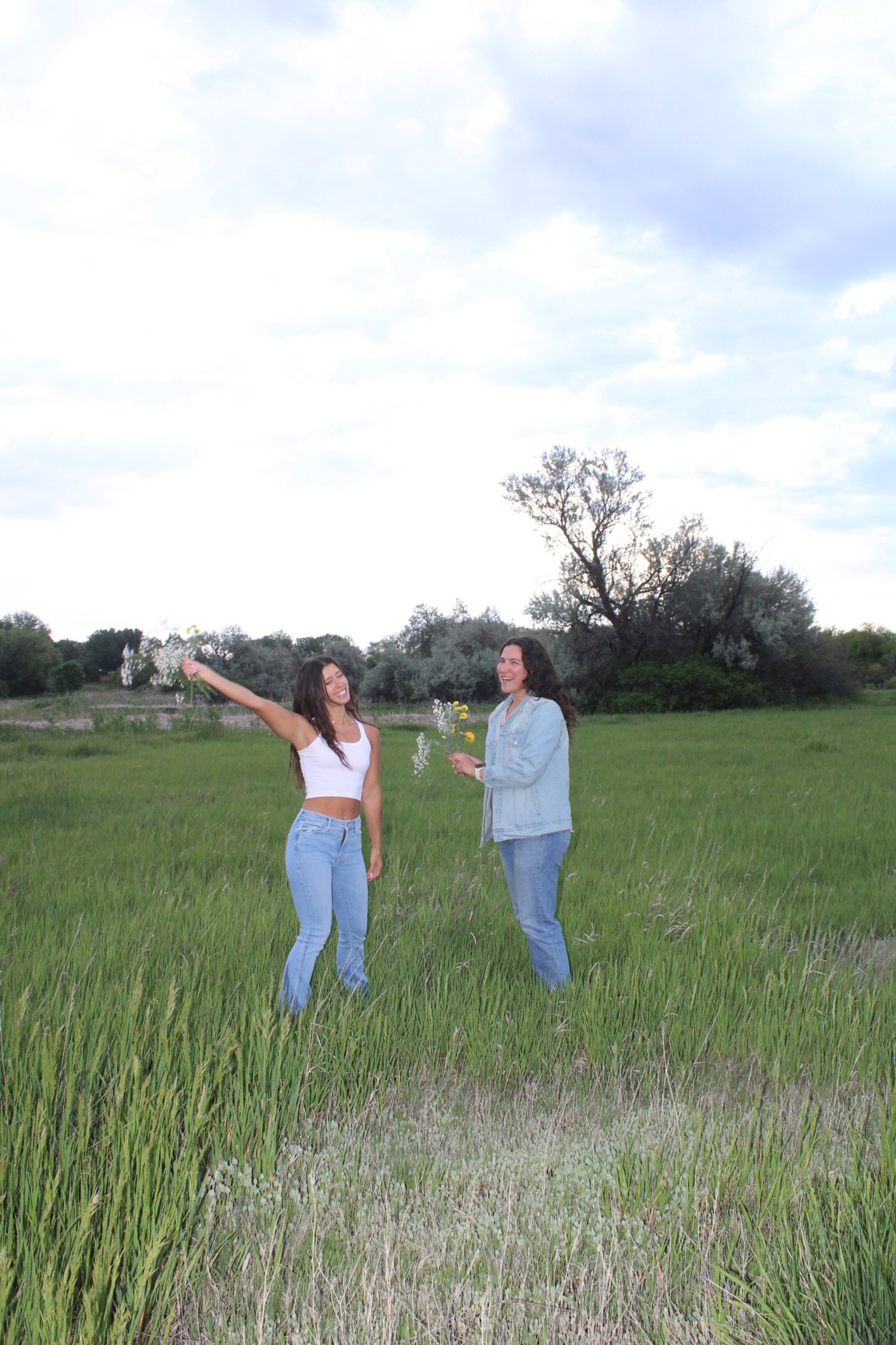 Two women standing in a grassy field, smiling, with one woman holding a small bouquet of wildflowers, and the other woman raising her arm, outdoors on a partly cloudy day.