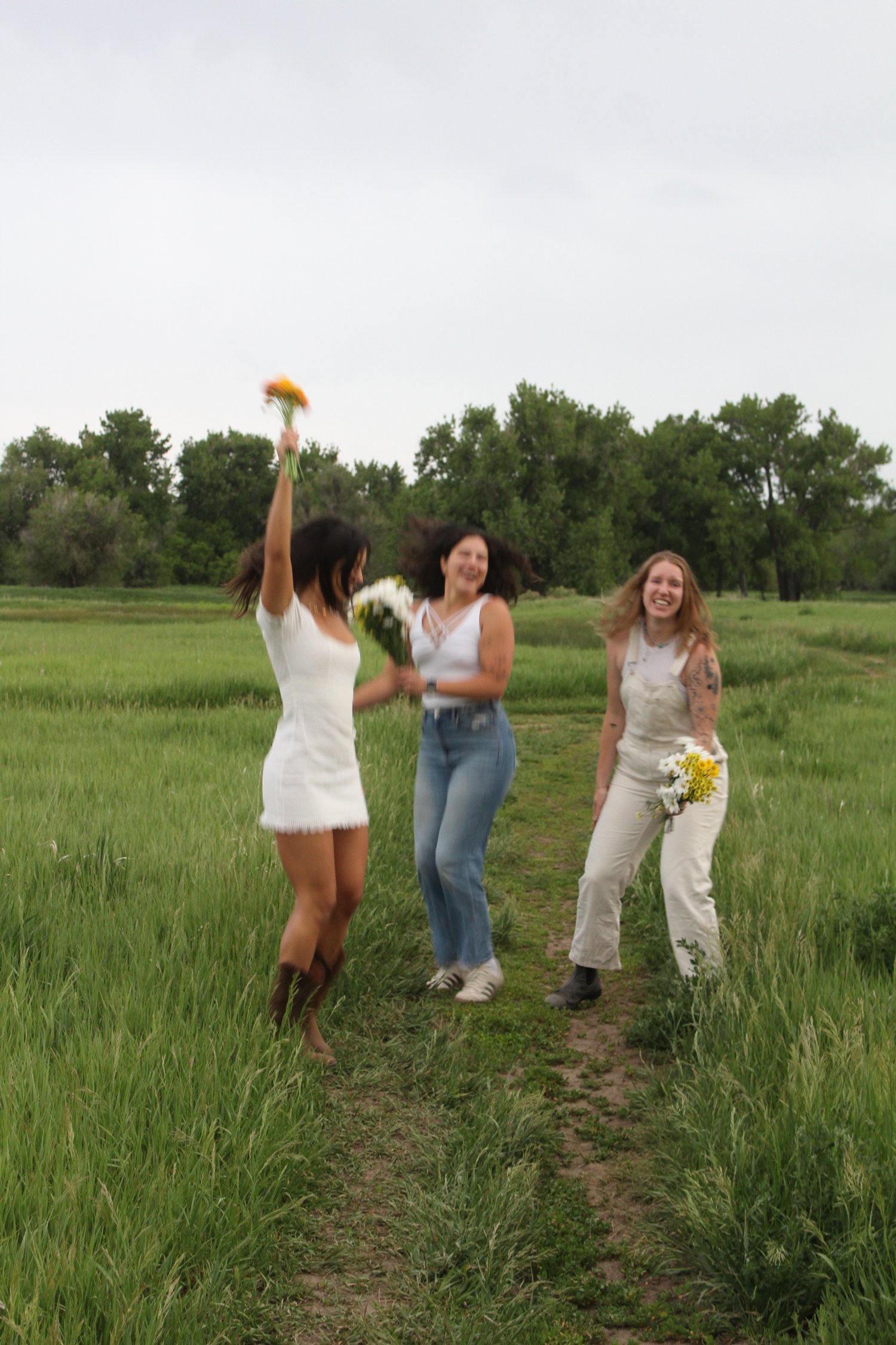Three women celebrating outdoors in a grassy field, holding flowers and jumping with joyful expressions on a cloudy day.