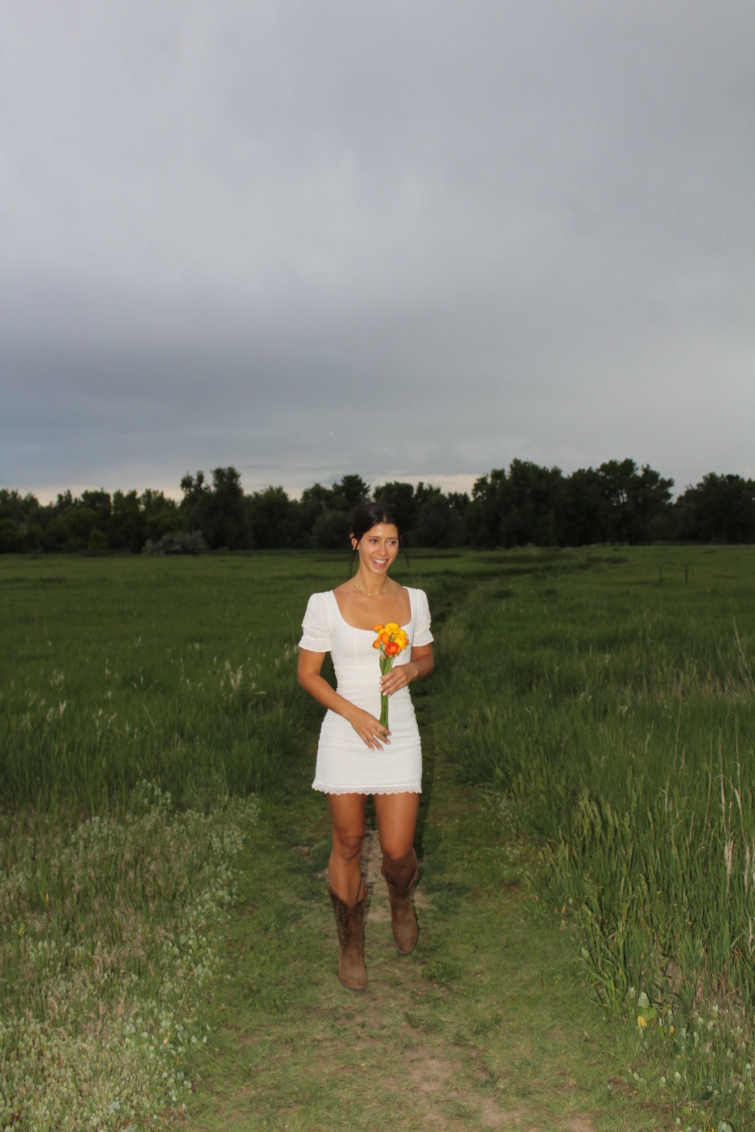 Woman walking on a grassy path through a field holding a bouquet of yellow and orange flowers, with a cloudy sky overhead.