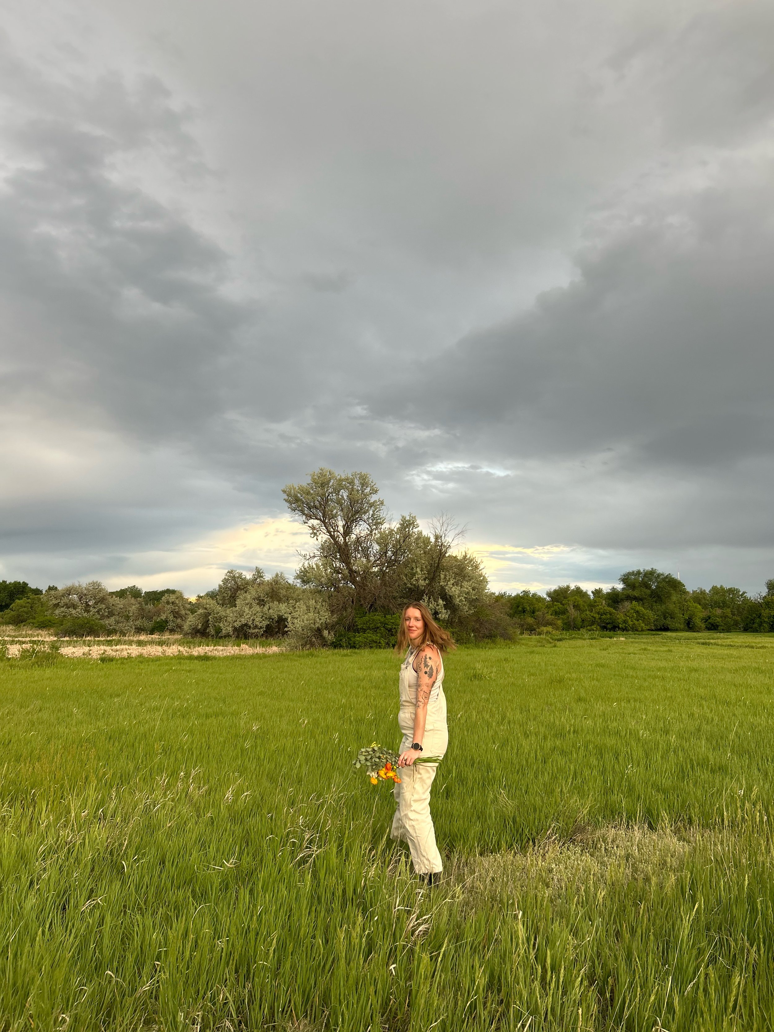A woman with tattoos on her arms and long hair standing in a lush green field, holding a small bouquet of flowers and looking at the camera, with overcast sky in the background.