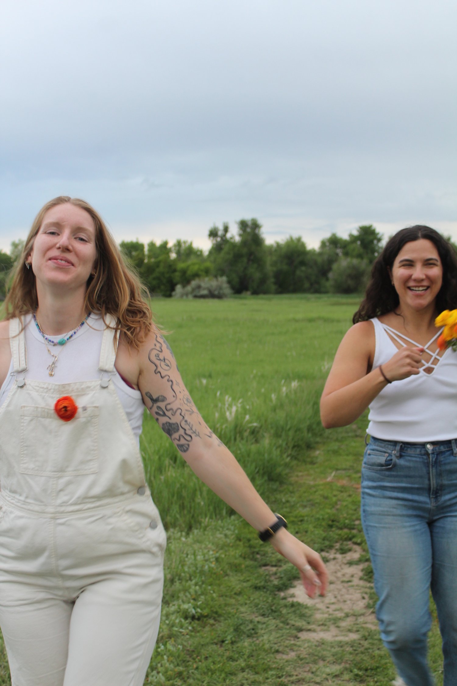 Two women walking outdoors in a grassy field, smiling and holding flowers, with trees and a cloudy sky in the background.
