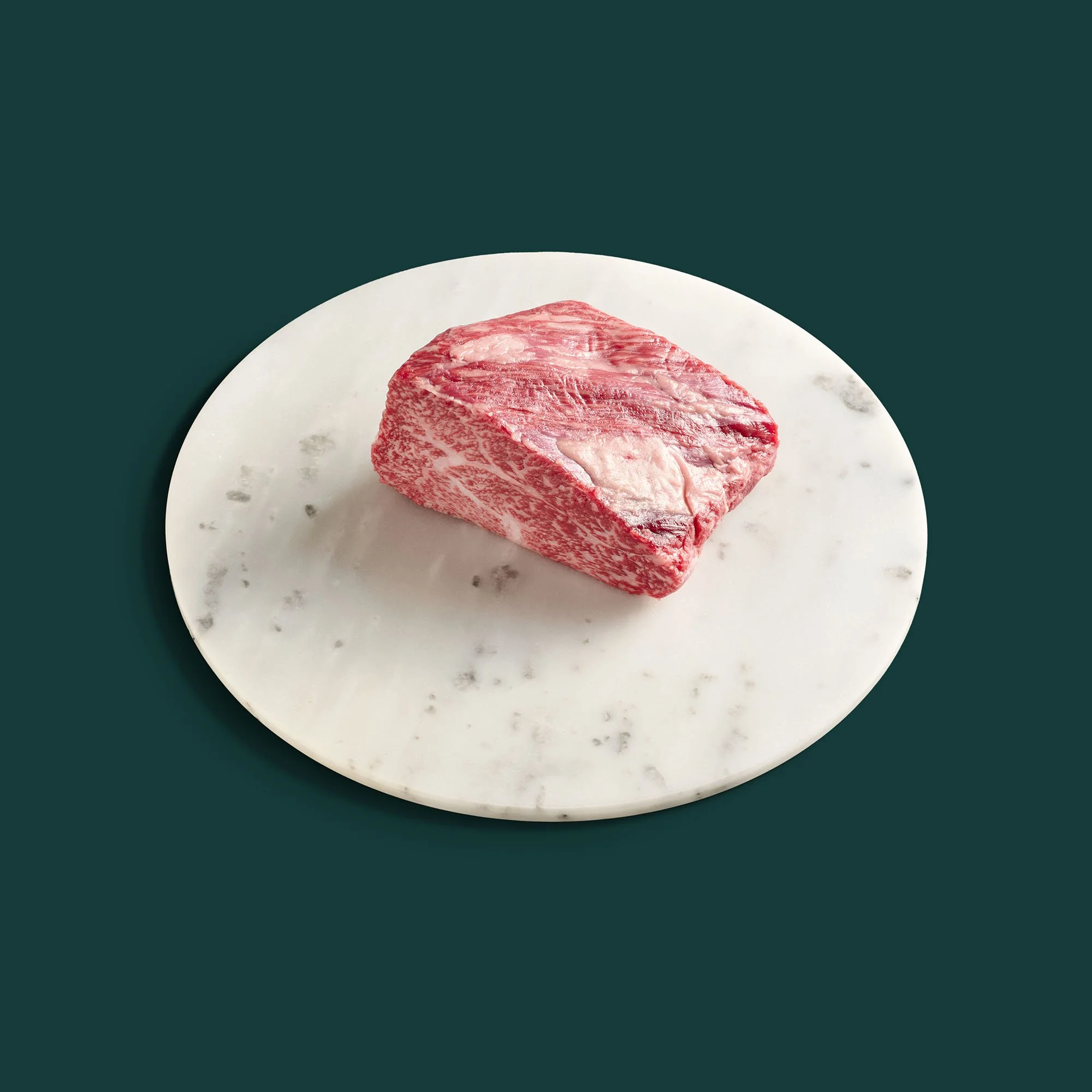 A single raw tri-tip steak with an angular shape and visible marbling sits off-centre on a ceramic plate, spotlighted from the right.