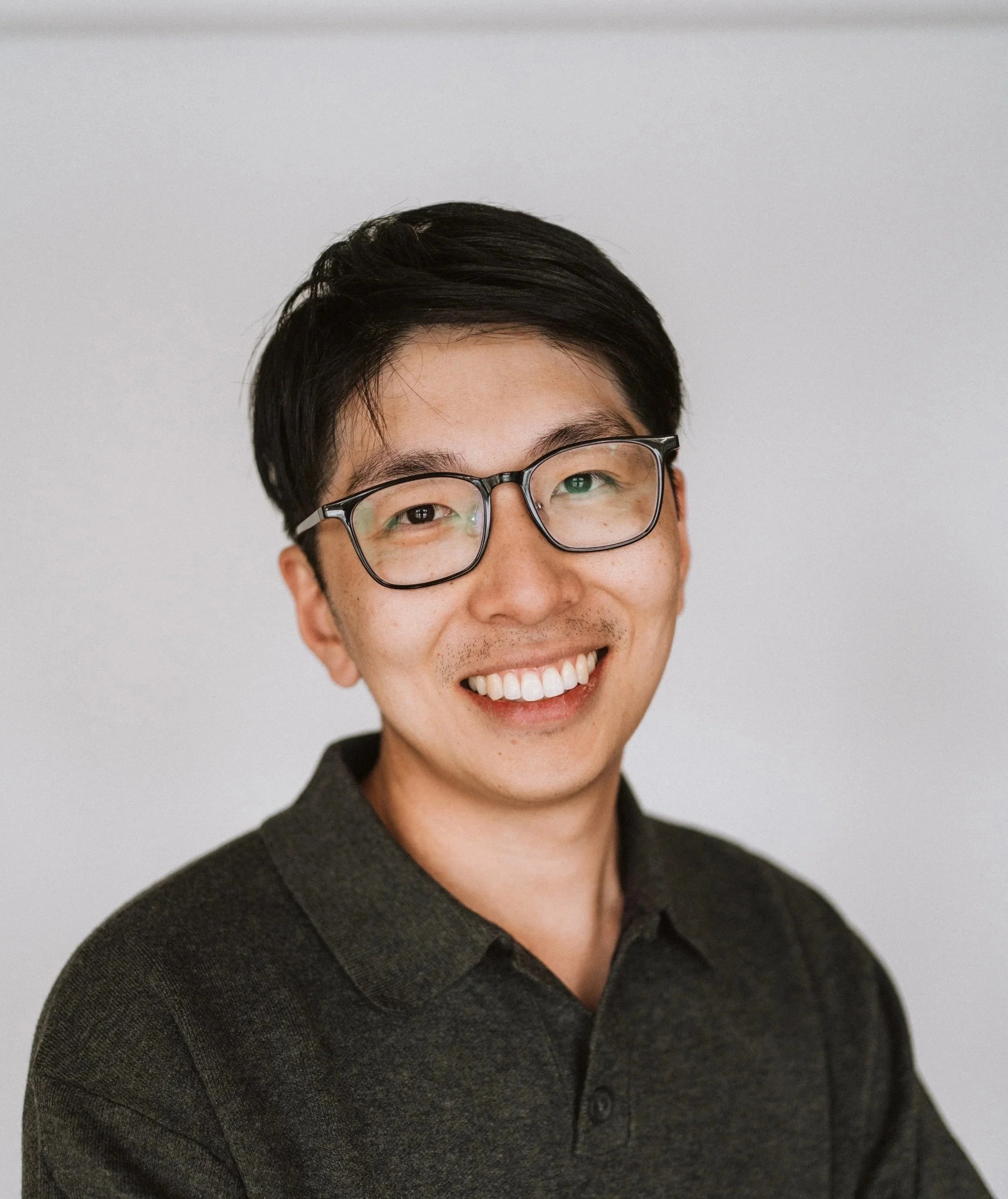 A smiling man with black hair, glasses, and a dark polo shirt against a plain light background.