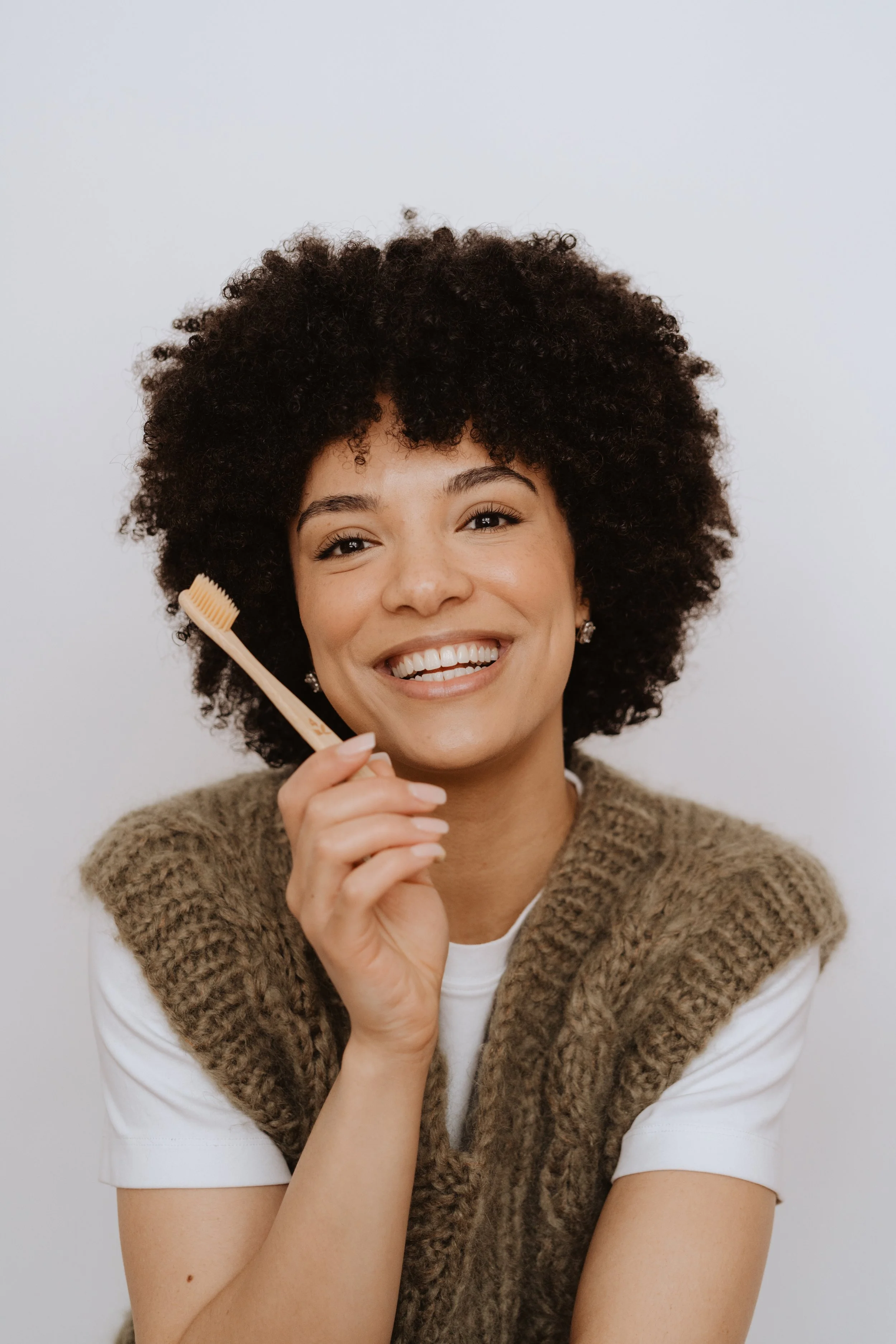 A woman with curly dark hair smiling at the camera, holding a beige toothbrush near her cheek, wearing a white shirt and a brown knit vest against a plain white background.