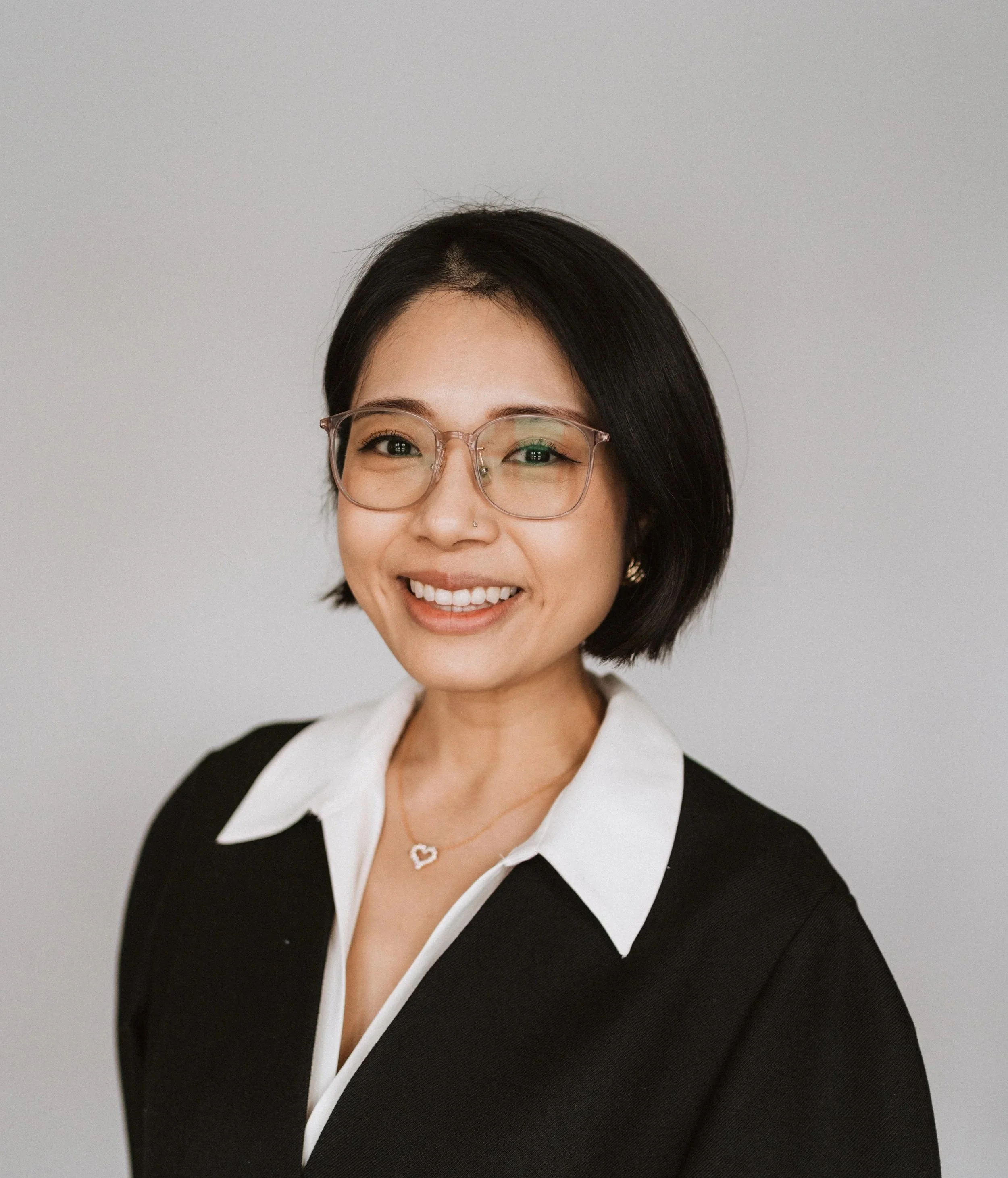A woman with short black hair, glasses, and a nose piercing, smiling and wearing a black top with a white collar, and a necklace with a heart-shaped pendant, against a plain light gray background.