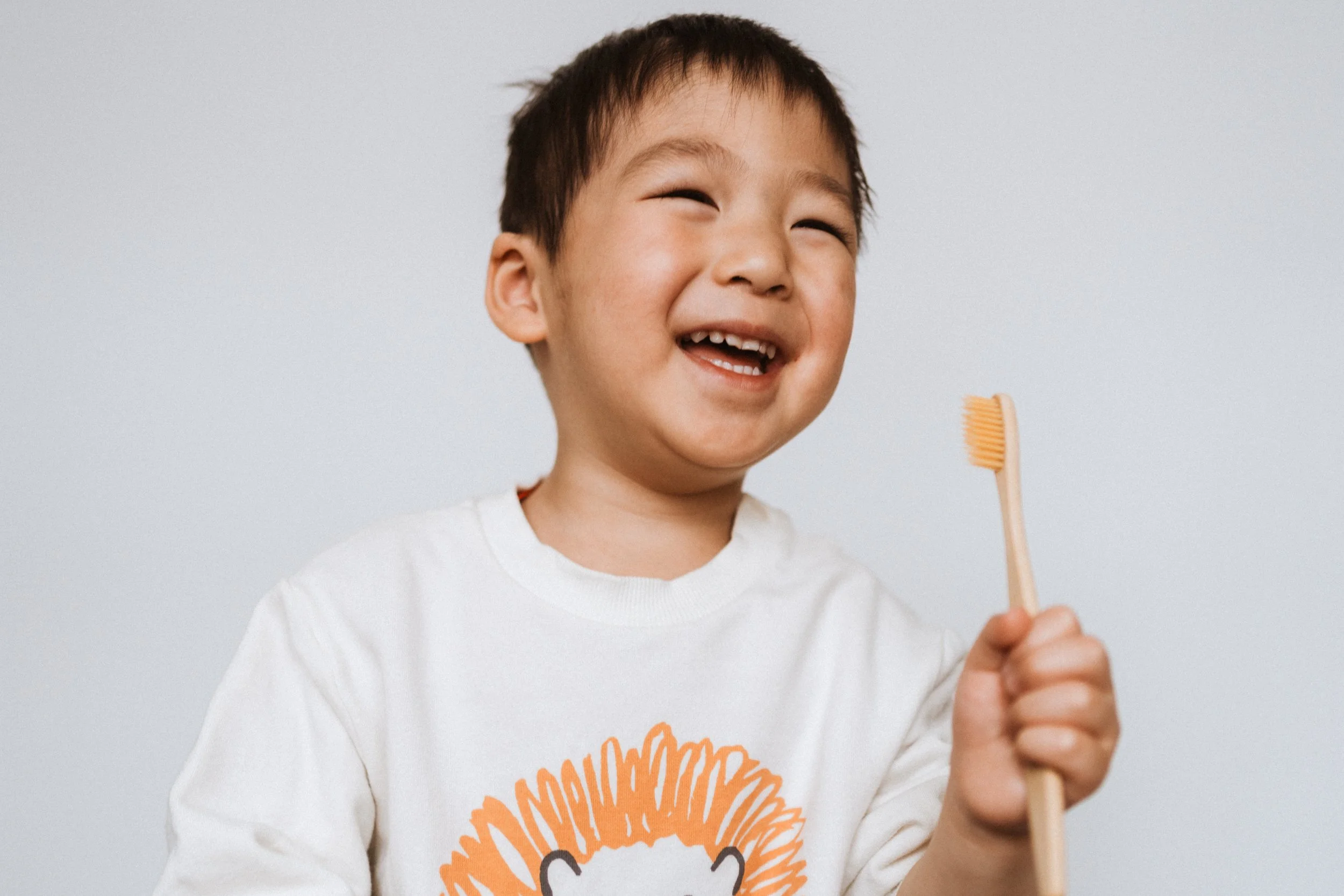 A young boy with short brown hair, smiling and holding a toothbrush, wearing a white shirt with an orange lion face design.