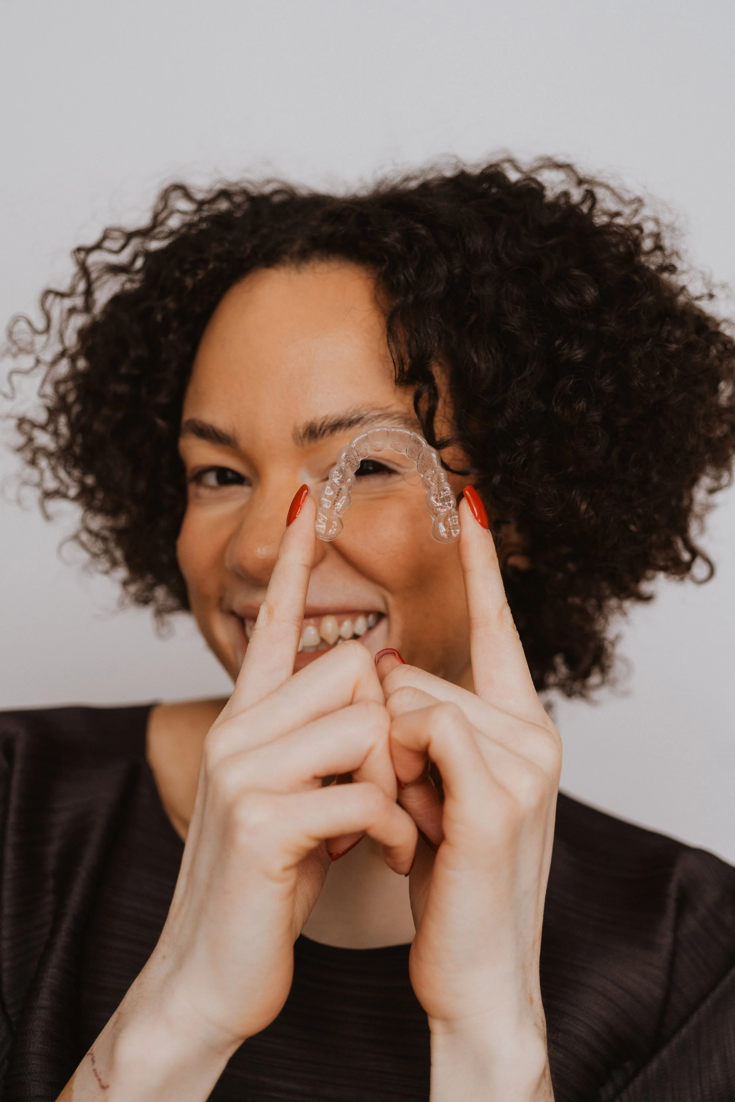 Woman with short curly hair holding an orthodontic retainer in front of her face, smiling.