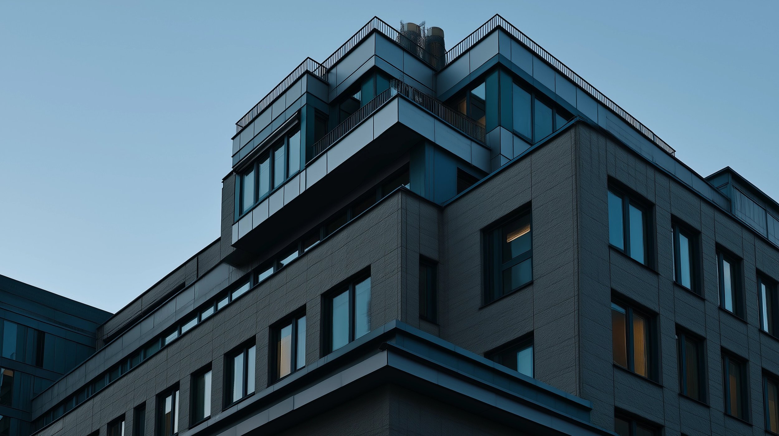Modern multi-story building with large glass windows and a rooftop terrace, set against a clear blue sky.