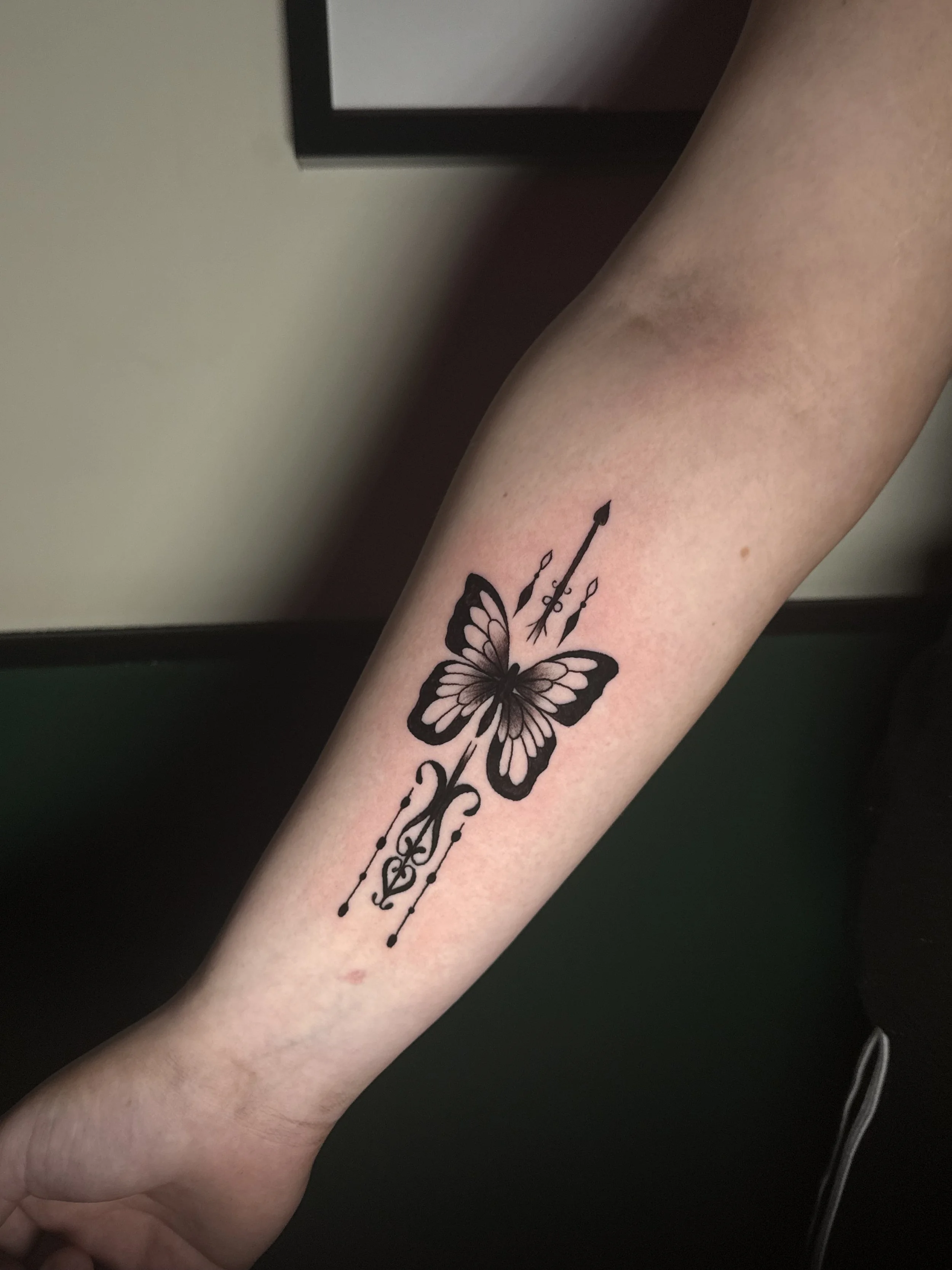 A person's forearm with a black butterfly and arrow tattoo.