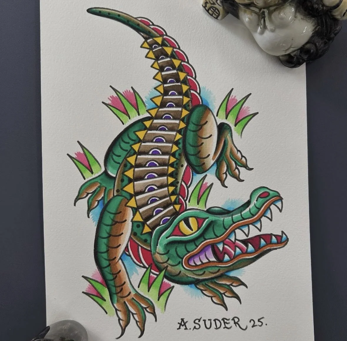 Colorful tattoo-style illustration of a crocodile with a long, armored back surrounded by pink and green grass, signed by A. Suder, 25.