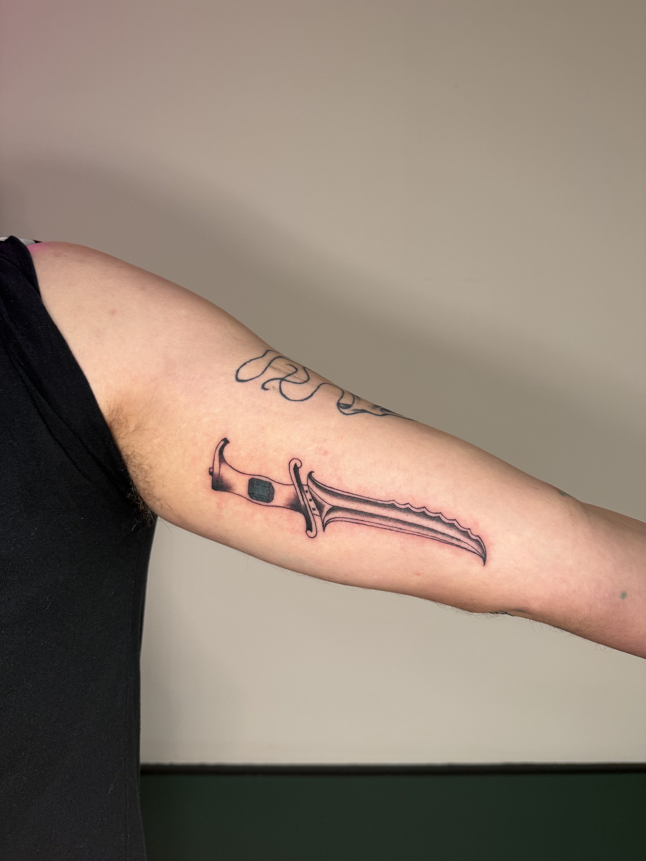 A person's upper arm with a tattoo of a sword and decorative elements.