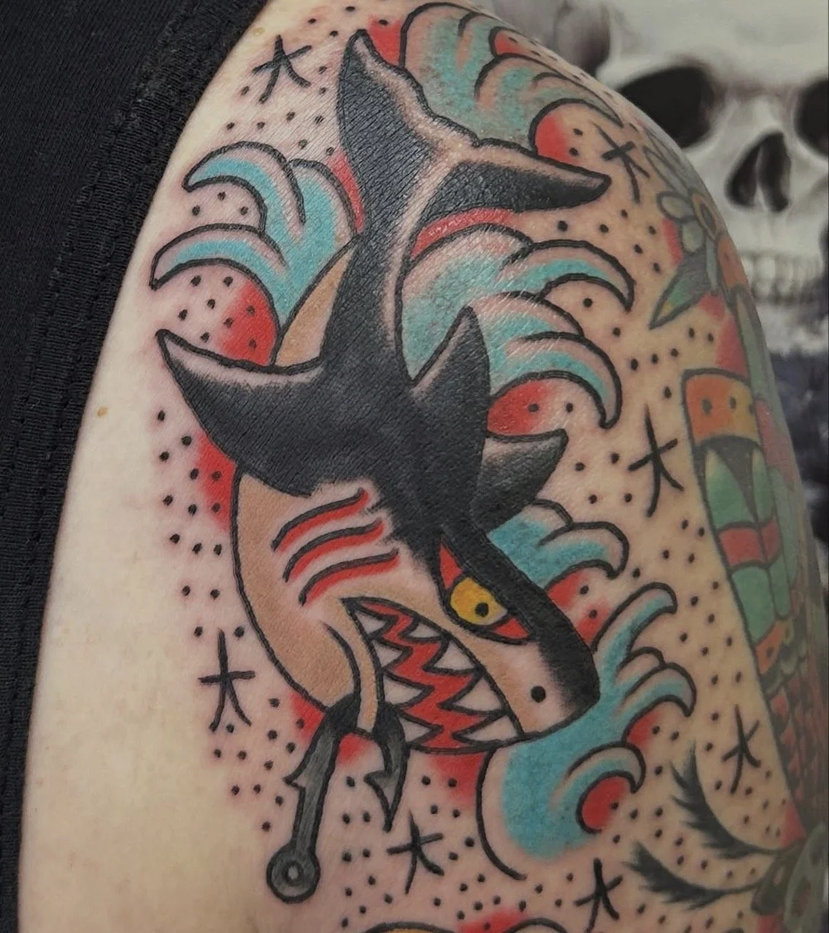 Tattoo of a black shark with yellow eye on an arm, surrounded by colorful waves, dots, and star-like shapes.
