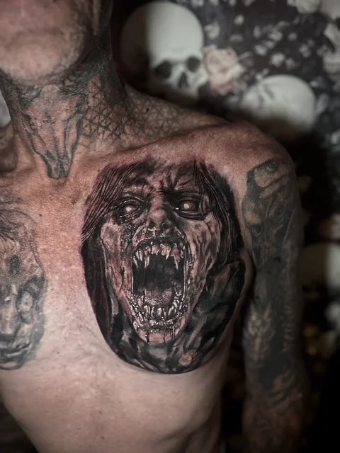 Tattoo of a screaming, ghoulish face on a person's chest, with skulls visible in the background.
