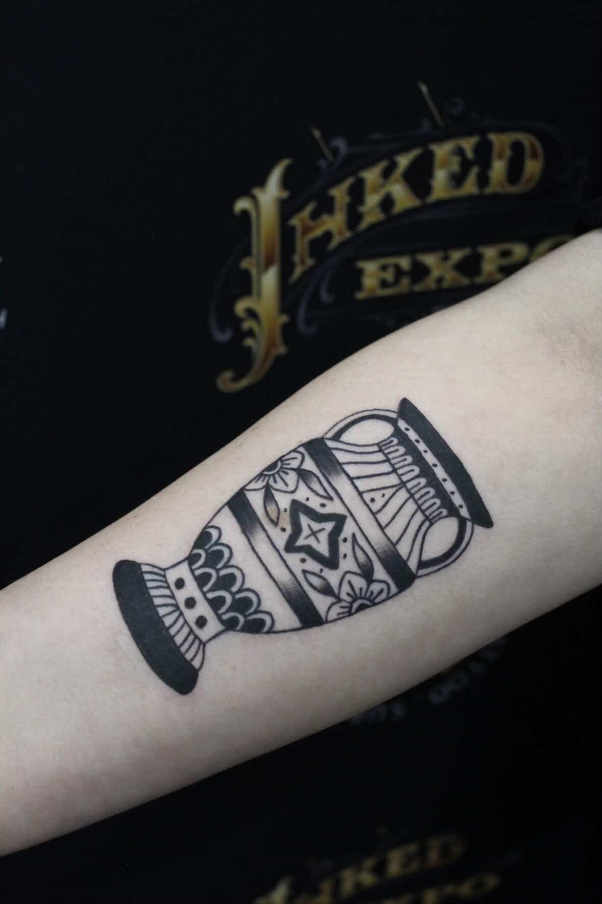 Black outline tattoo of a vase with decorative patterns on a person's forearm.